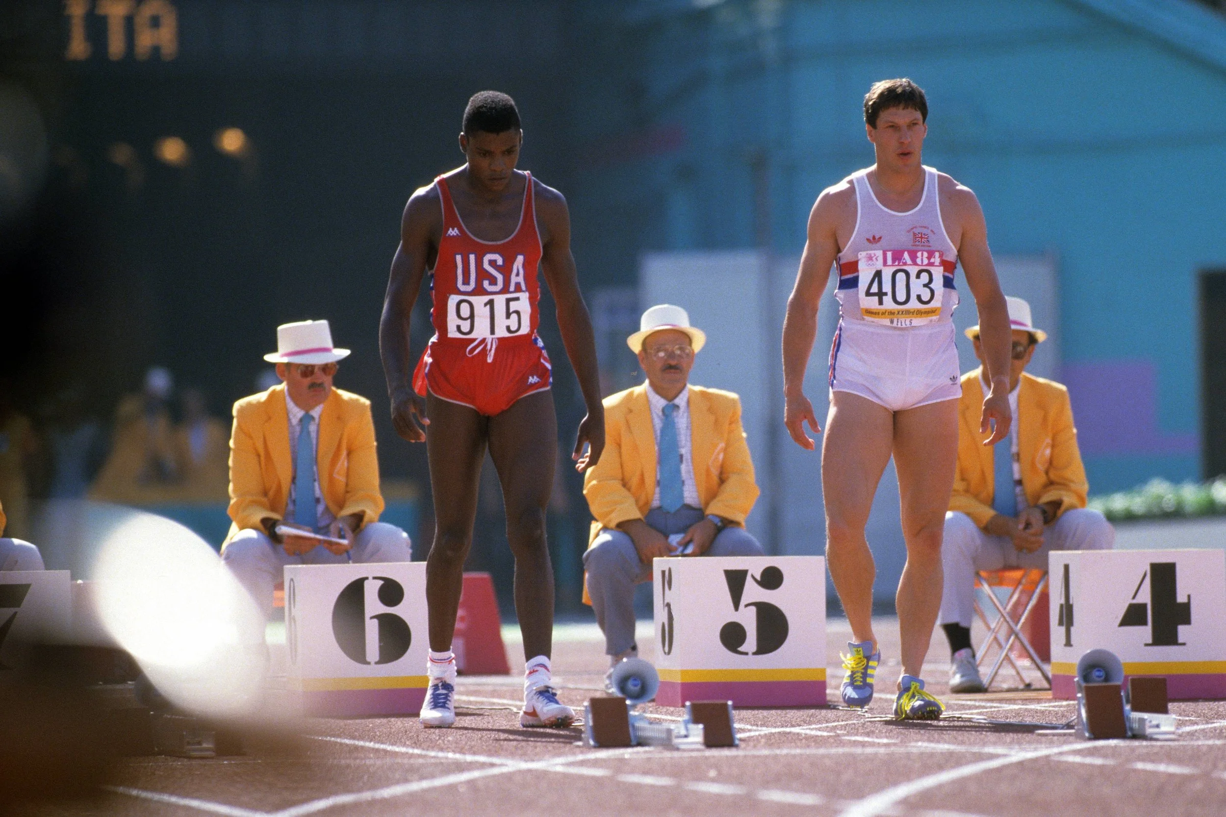 Two male track athletes at a starting line, preparing for a race, with officials sitting behind them and holding clipboards. One athlete wears a red jersey with 'USA' and the number 915, the other wears a white jersey with red and blue accents and the number 403. They are on a running track in an outdoor stadium.