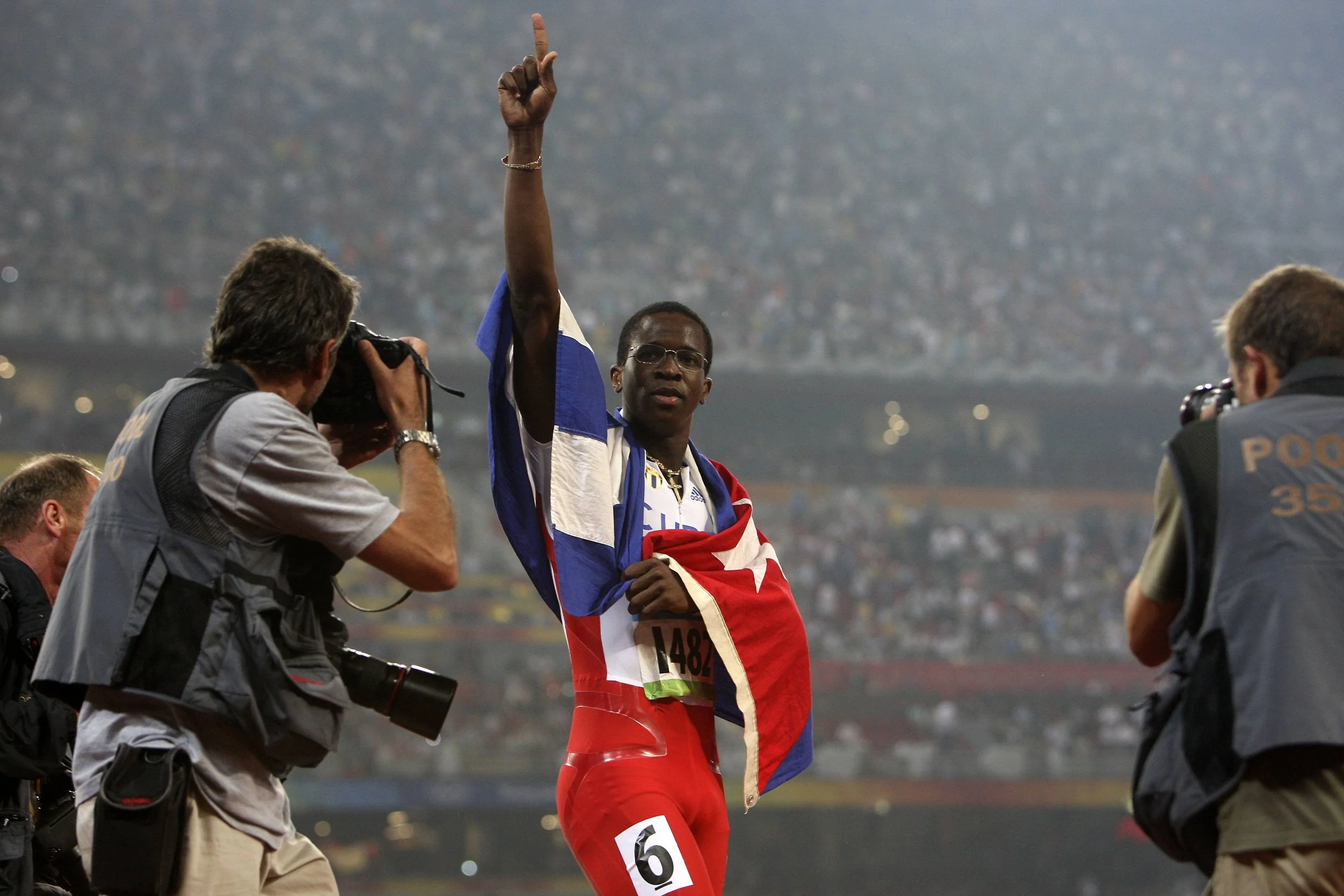 A man wearing a British flag scarf and red sports gear, holding a race bib with the number 6, raising his right index finger in the air at a stadium event with photographers capturing his image.