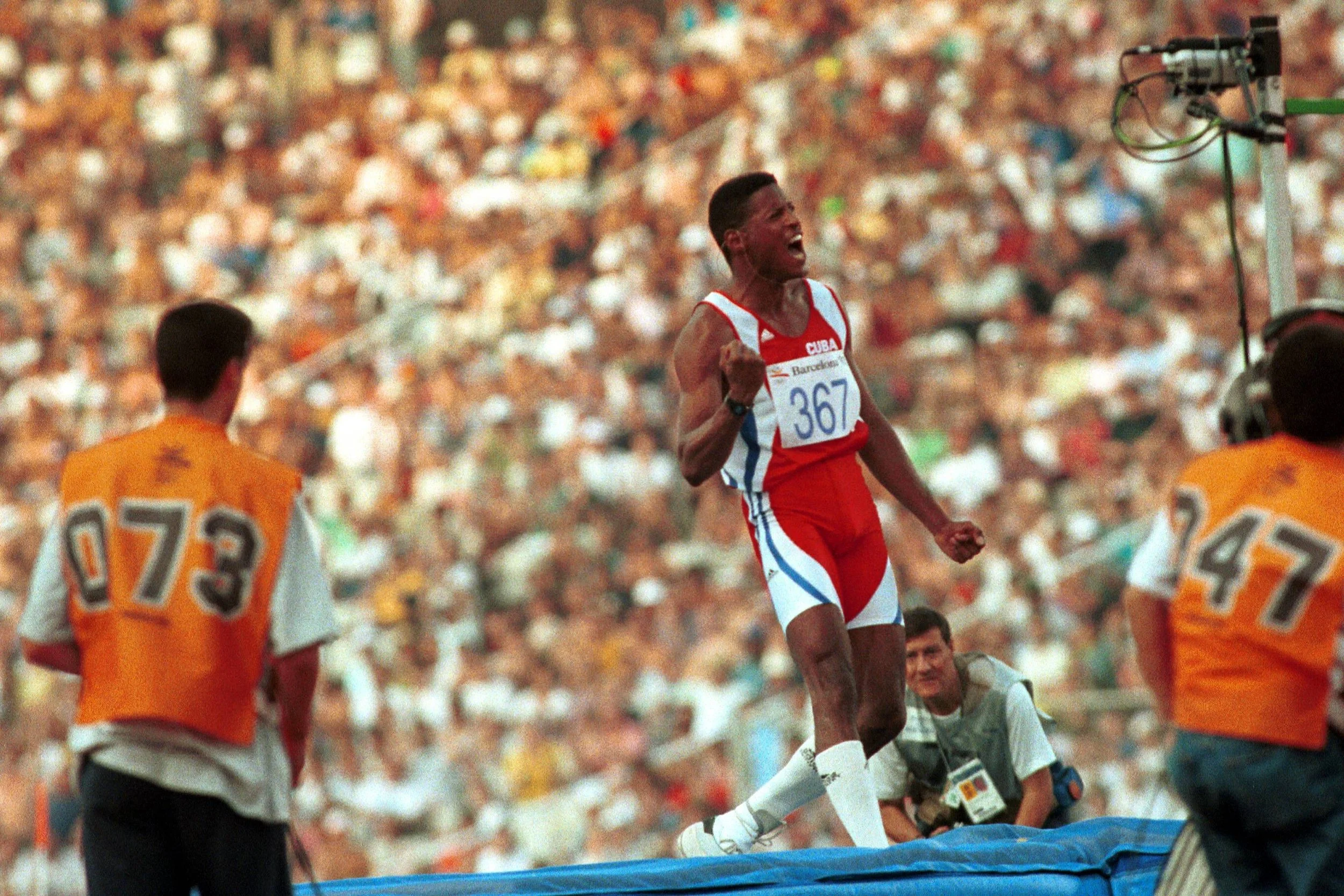 A male athlete in a red and white uniform celebrating during a track and field event, with a large crowd in the background and officials or helpers nearby.