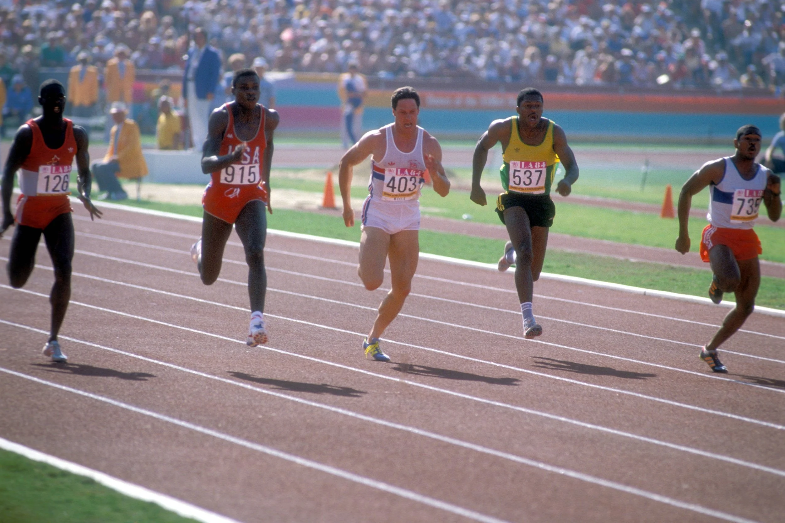 Several runners competing in a track race outdoors on a sunny day, with a large crowd of spectators in the background.