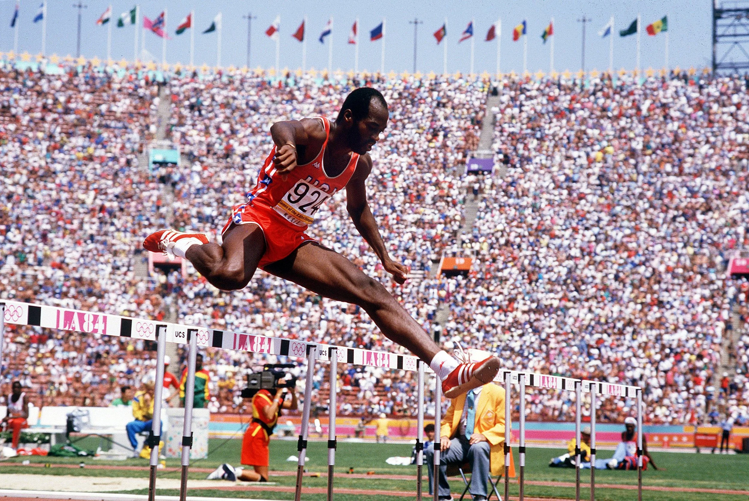A male athlete in a red uniform with the number 924 competes in a hurdles race at the 1984 Los Angeles Olympics, with a large crowd and multiple flags in the background.