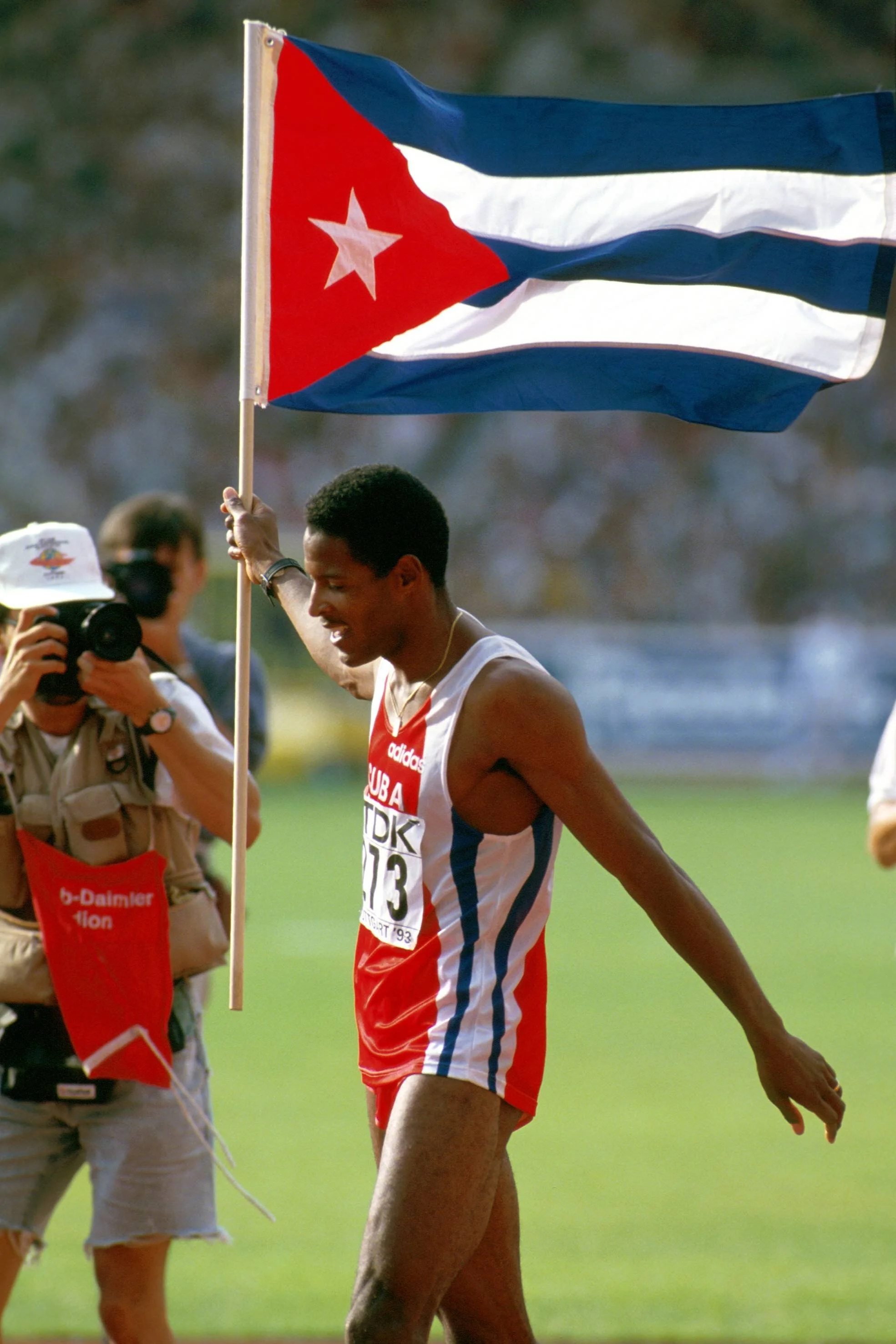 A male athlete holding a Cuban flag after a race, wearing a red, white, and blue athletic uniform, with a photographer and a person with a cap nearby.