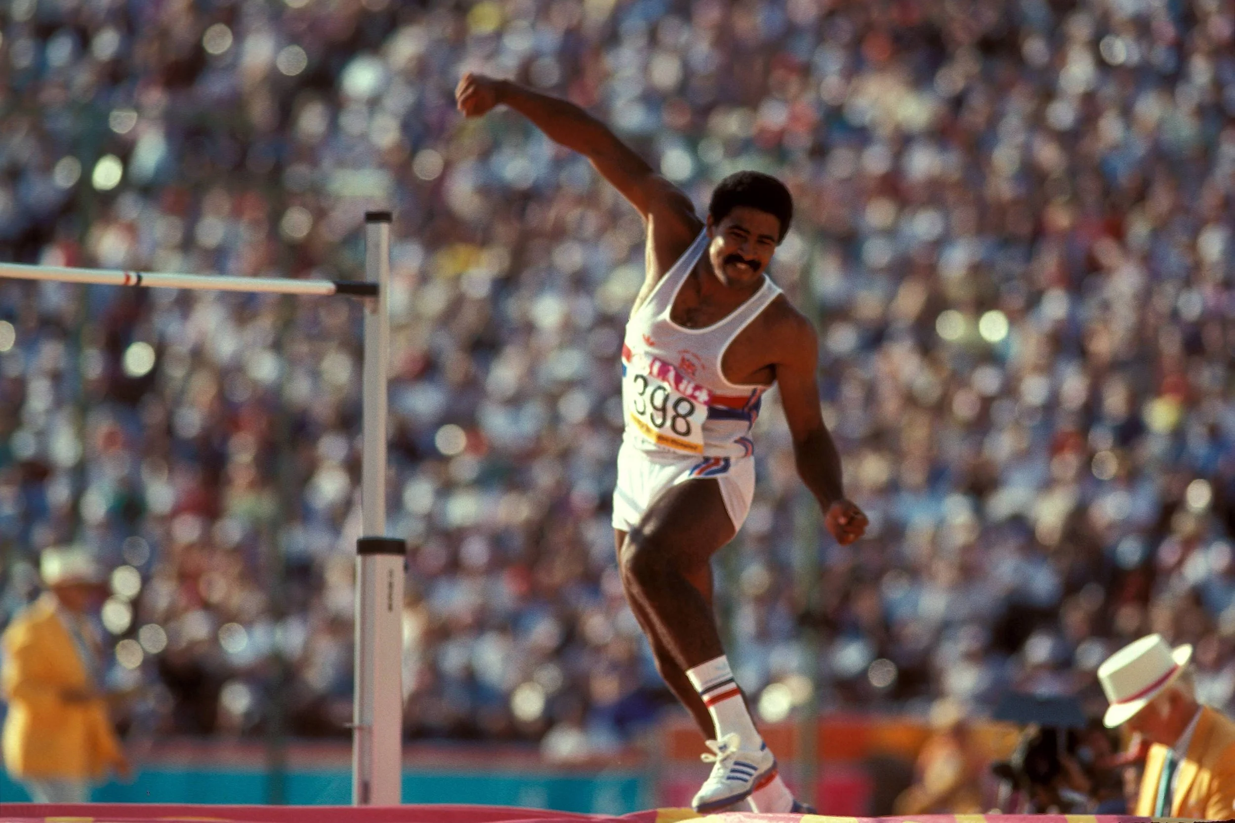 A male track athlete crossing the hurdle during a race at a stadium filled with spectators.