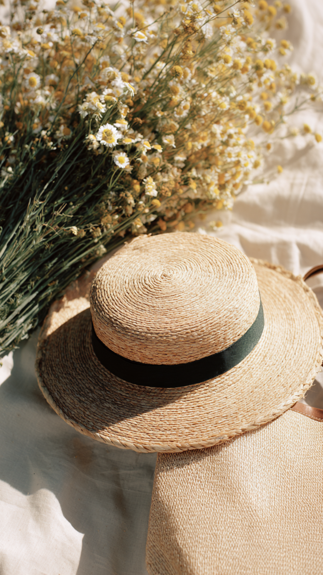 A straw sun hat with a black ribbon, a bouquet of small white and yellow flowers, and a cream-colored textured fabric.