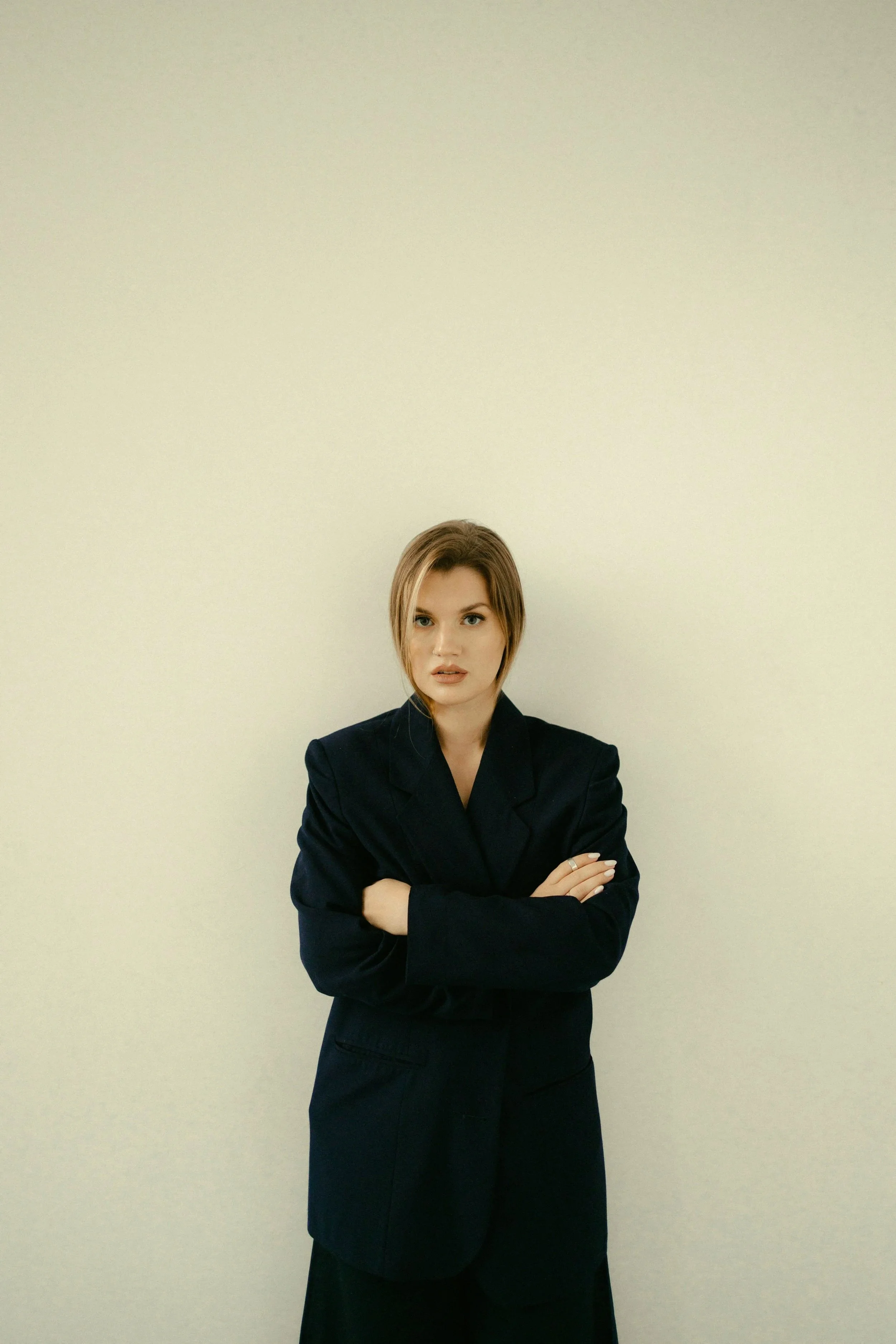 A woman with blonde hair, wearing a black suit, standing against a plain off-white wall, with arms crossed and a serious expression.