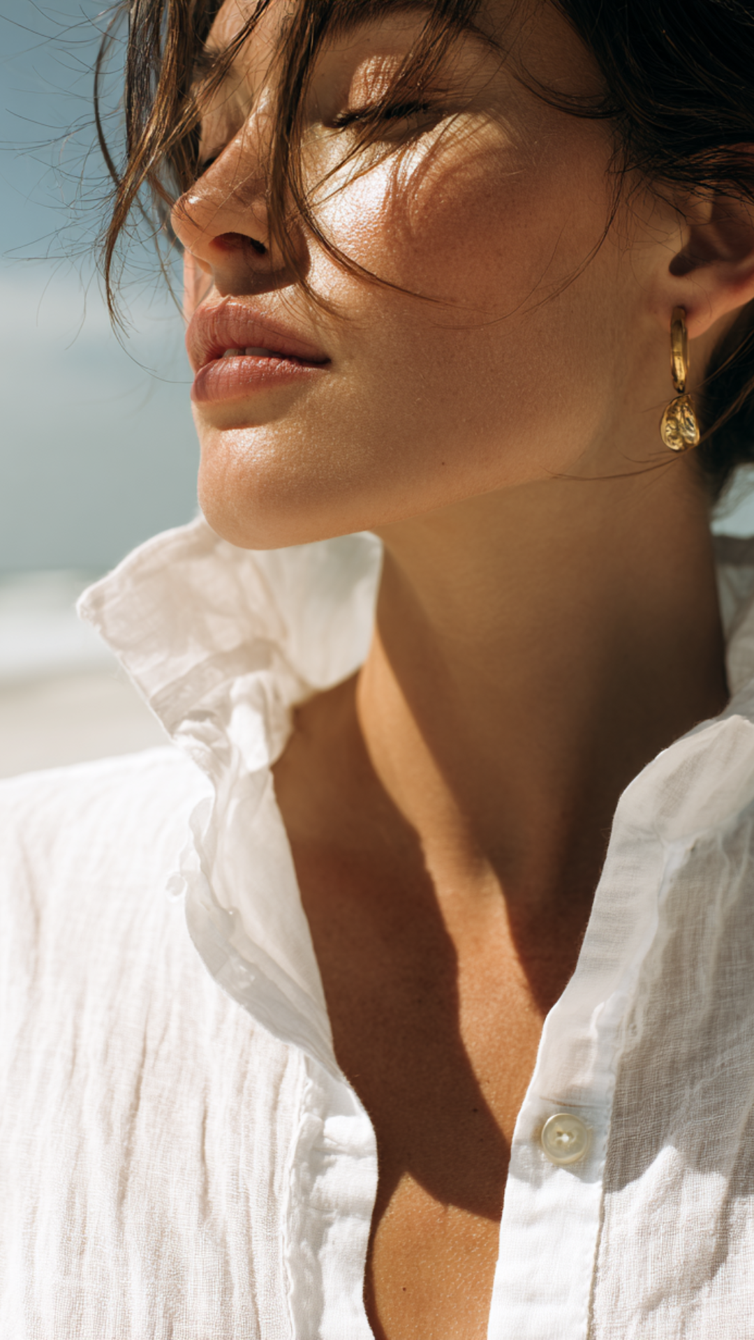 Close-up of a woman with tousled hair, wearing gold earrings, a white button-up shirt, with sunlight on her face and closed eyes.