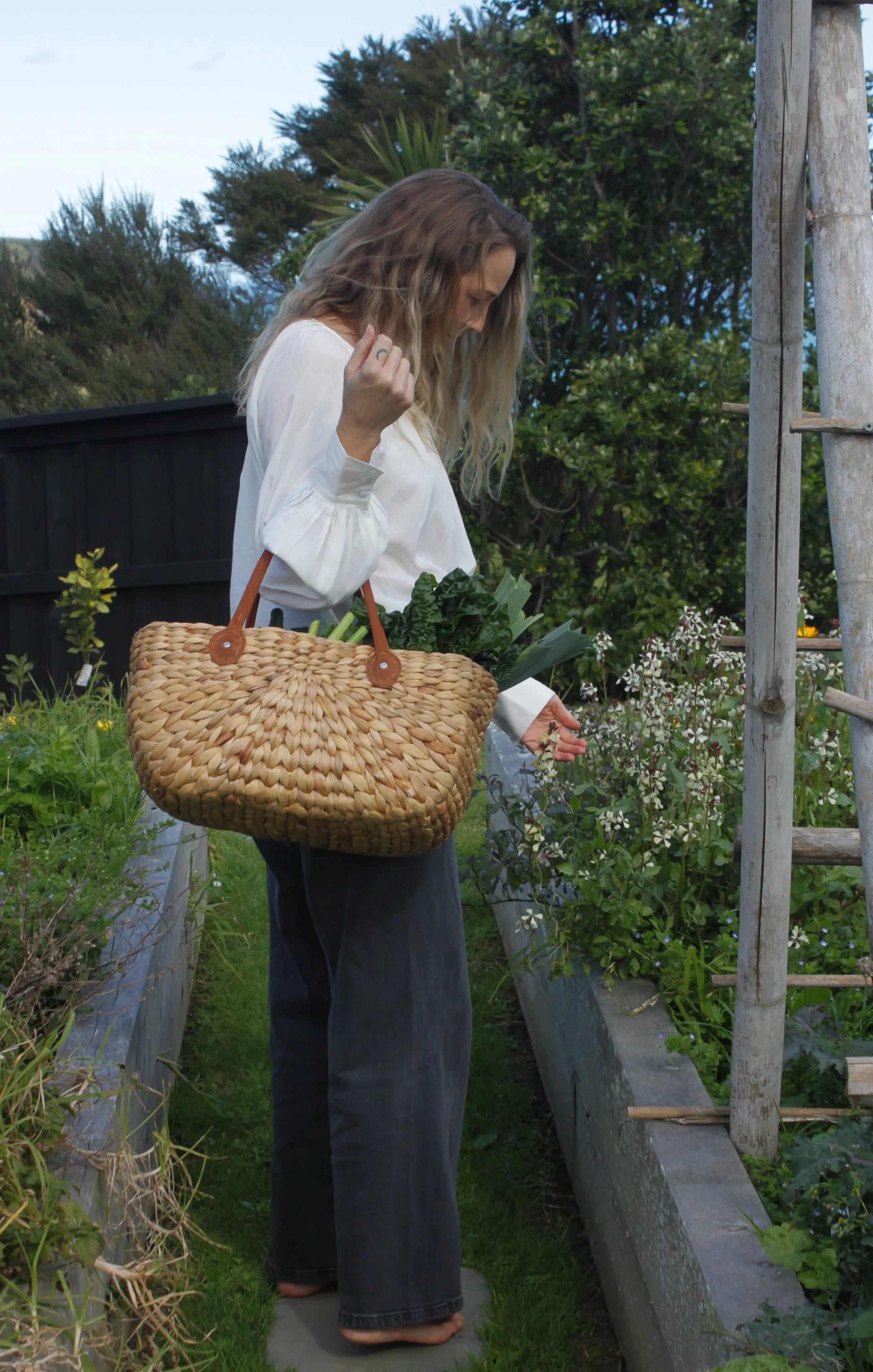 A woman with long wavy hair wearing a white blouse and dark jeans is standing barefoot in a garden or farm, holding a large woven basket filled with leafy greens. She appears to be inspecting or selecting plants along a garden bed, with trees and a blue sky in the background.