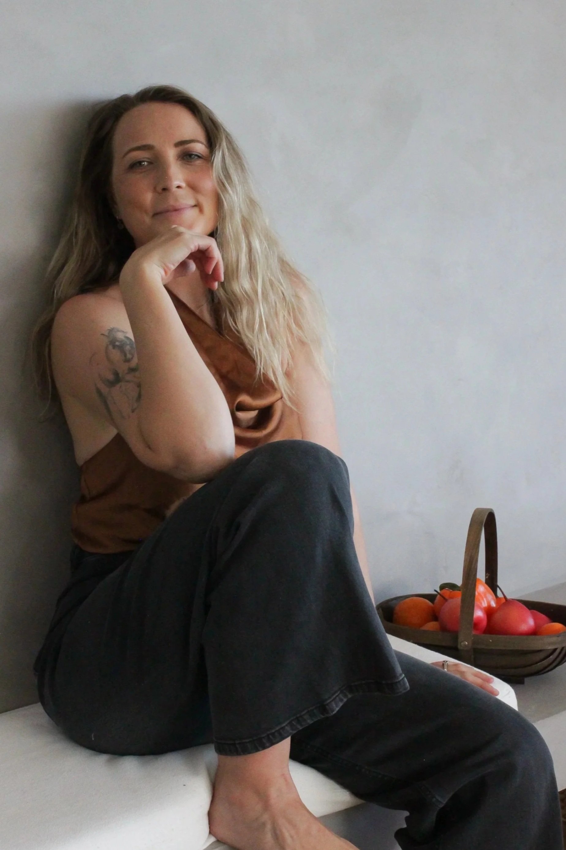 A woman with long, wavy, blonde hair, sitting against a plain wall, smiling gently. She has a tattoo of a tiger on her upper arm and is wearing a brown top and black pants. There is a basket of red apples on a table beside her.