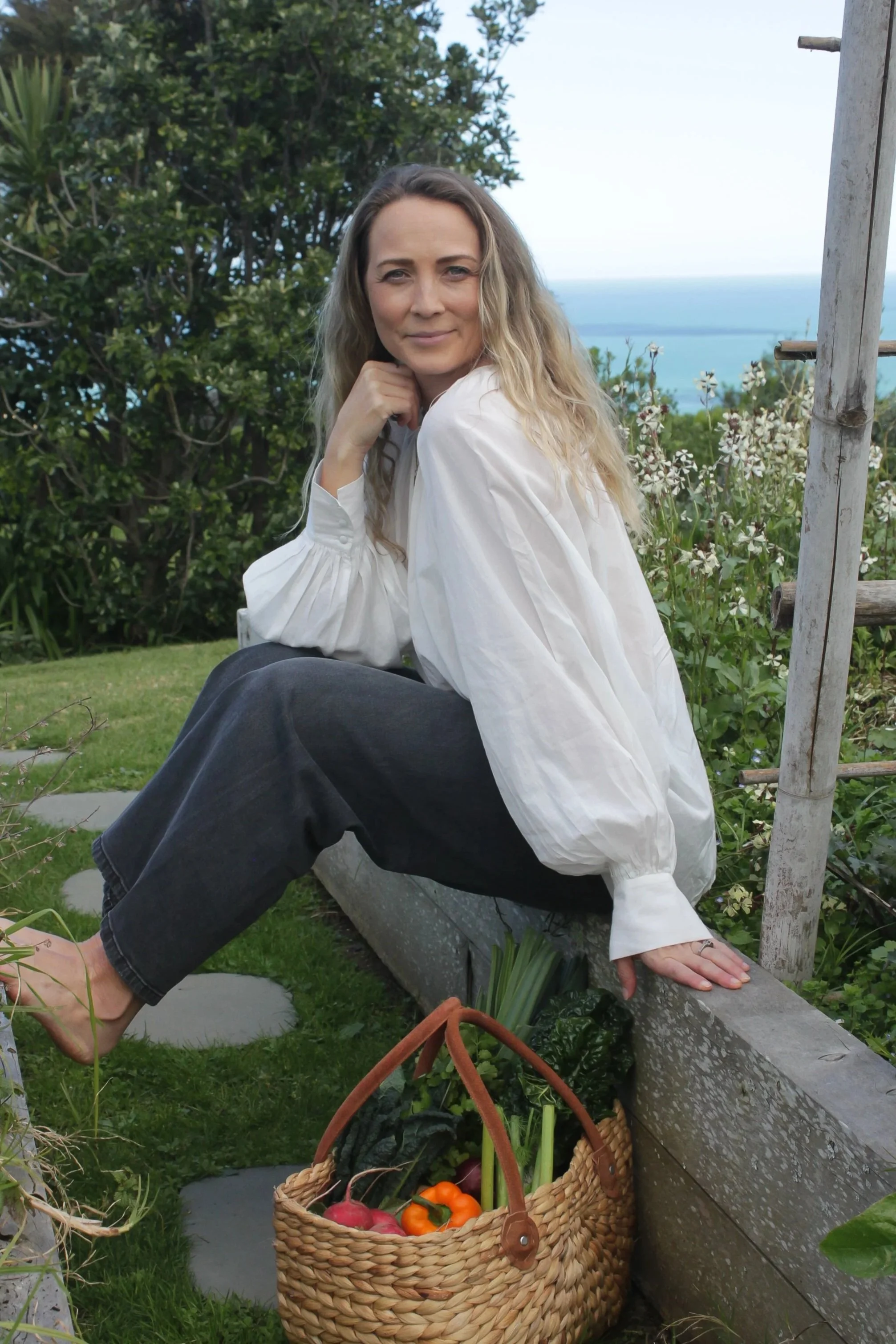 A woman with long wavy blonde hair sitting outdoors on a garden bench, wearing a loose white blouse and dark pants, with one hand resting on her knee and the other on the bench, a wicker basket filled with colorful vegetables like radishes, bell peppers, and carrots on the ground beside her, greenery and trees in the background, and the ocean visible in the distance.