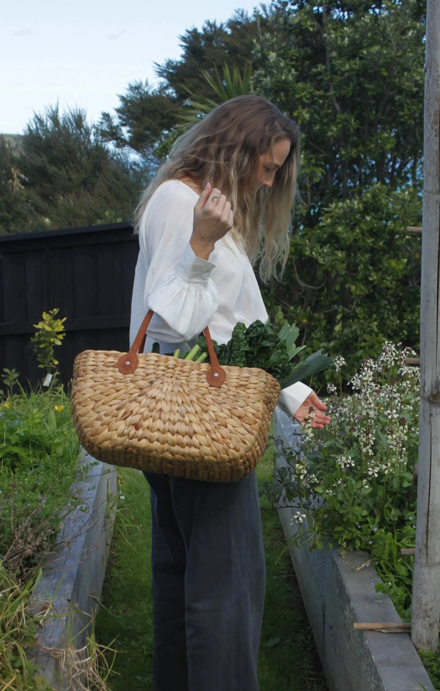 A woman with long wavy hair, wearing a white top and dark pants, stands in a garden, holding a large woven basket filled with leafy greens and vegetables, while inspecting plants.