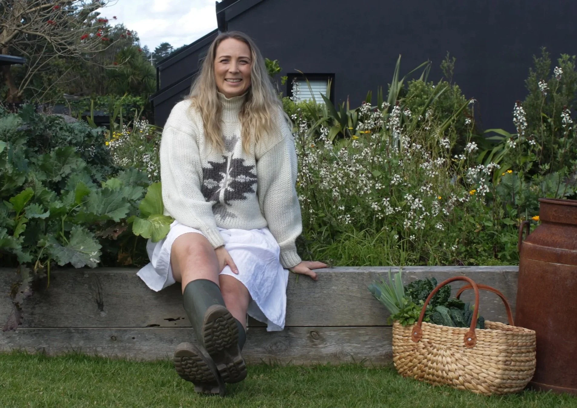 A woman sitting on a wooden garden border, smiling, wearing a white sweater with a tree design, a white skirt, and rubber boots, surrounded by lush greenery and flowers, with a black building in the background.