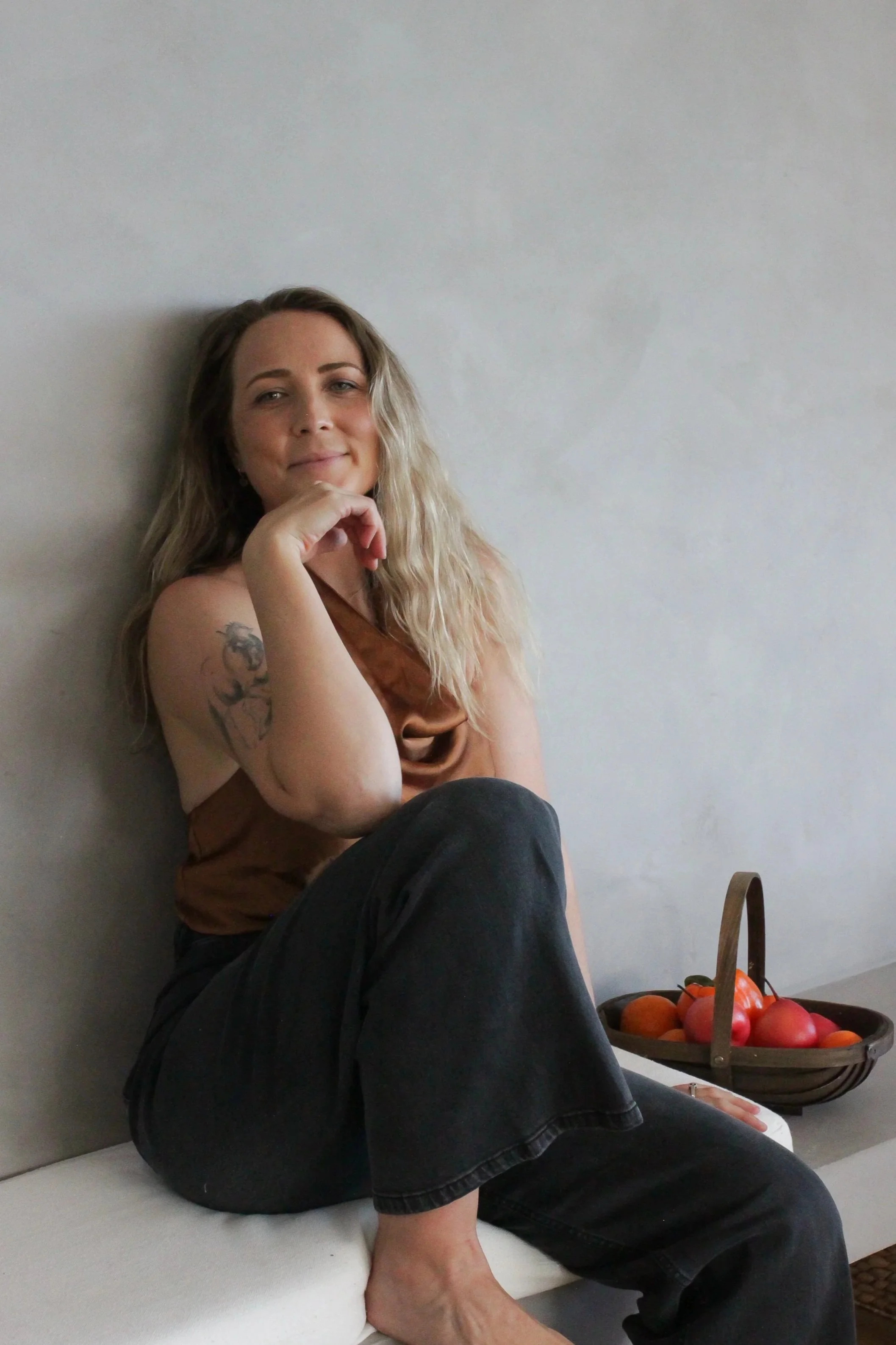 A woman with long blonde hair, wearing a brown top and black jeans, sitting on a white bench against a plain wall, resting her chin on her hand, with a basket of red apples on the floor beside her.