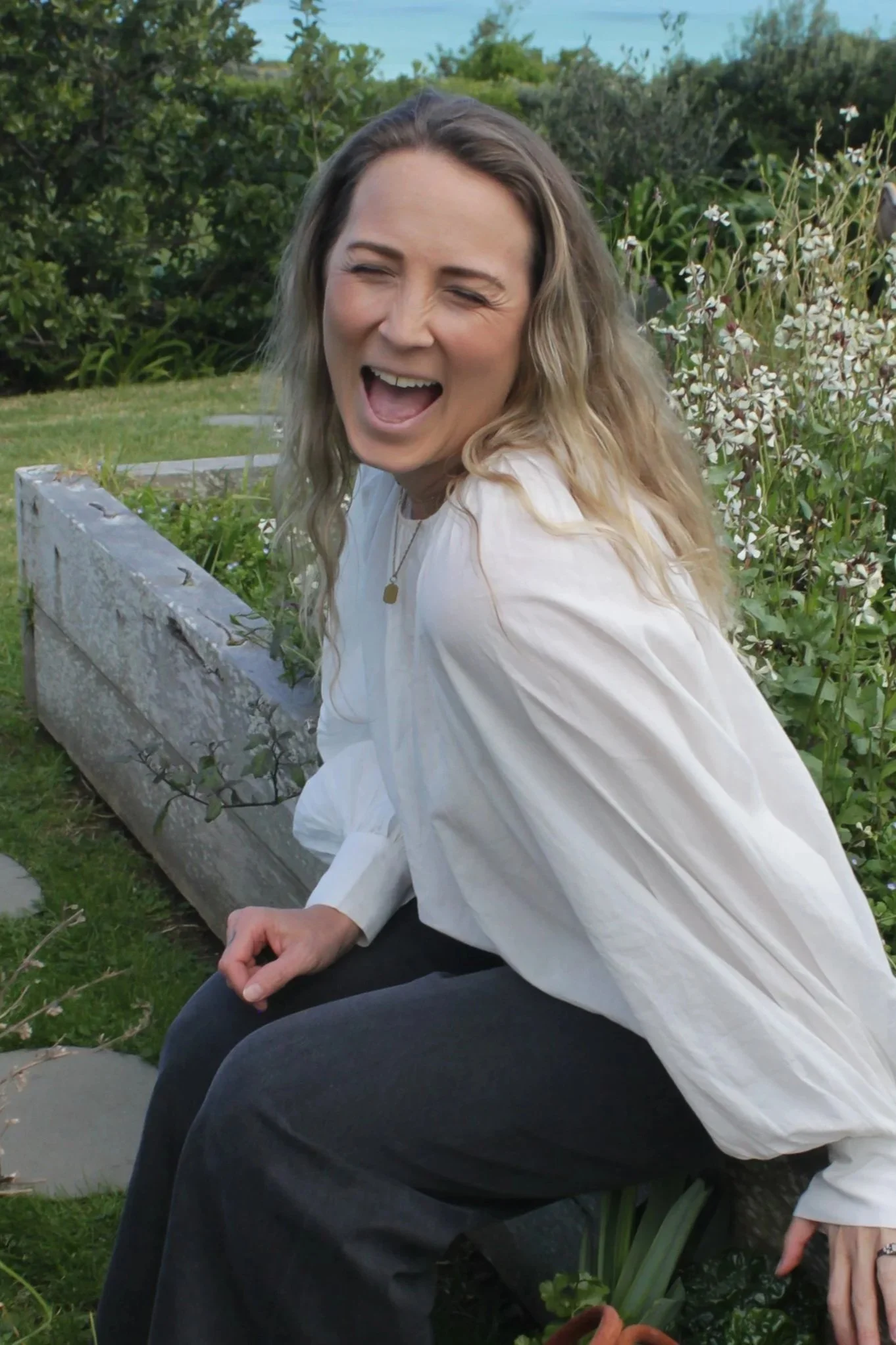A woman with wavy blonde hair, laughing or smiling, sitting outdoors in a garden with greenery, white flowers, and a weathered wooden planter.