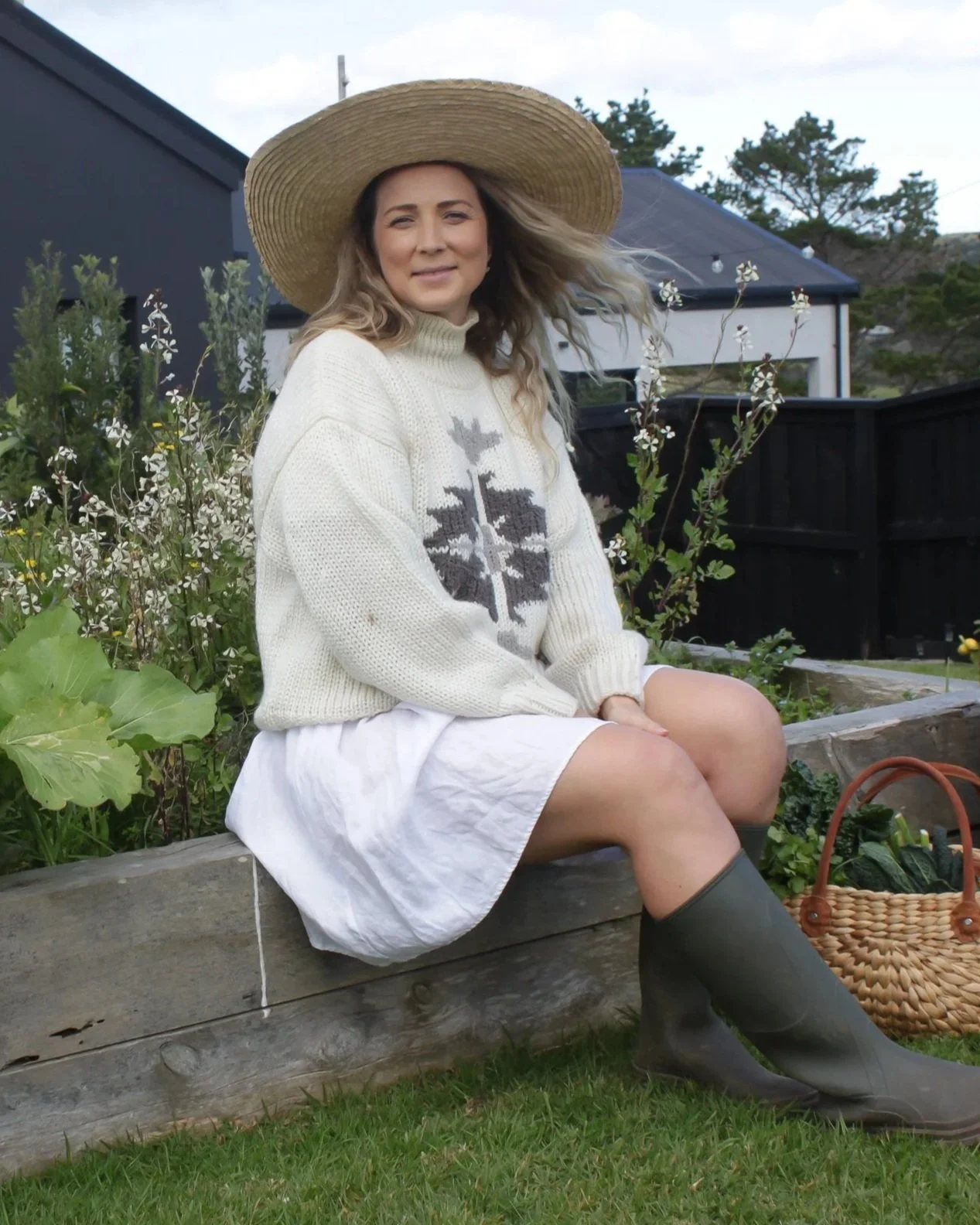 A woman sitting on a garden bench, wearing a large sun hat, cream sweater with a pattern, white skirt, and rain boots. She is surrounded by plants and flowers with a house in the background.