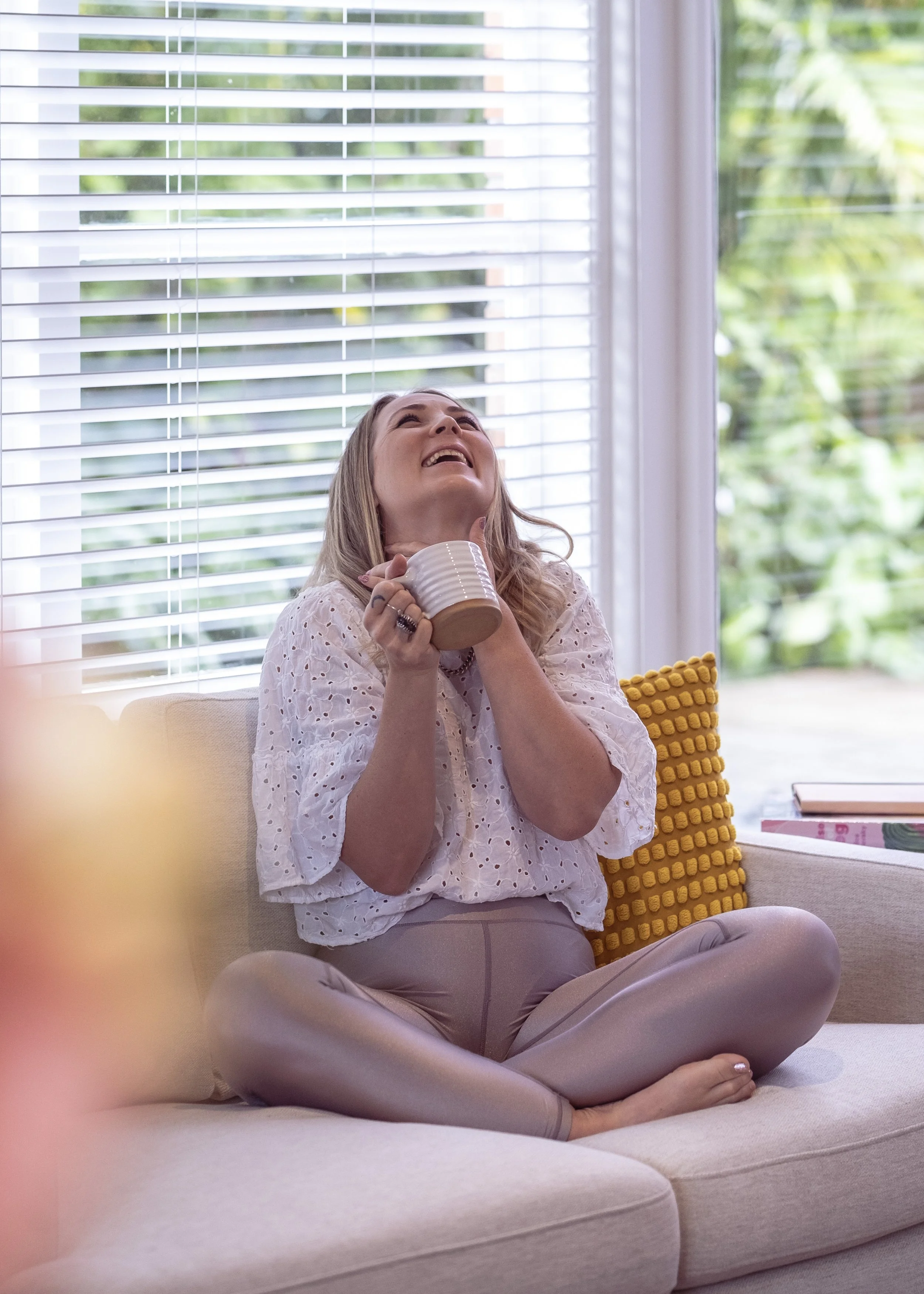 A woman sitting cross-legged on a beige couch, holding a mug, smiling, with her head tilted back near a window with blinds and green foliage outside.