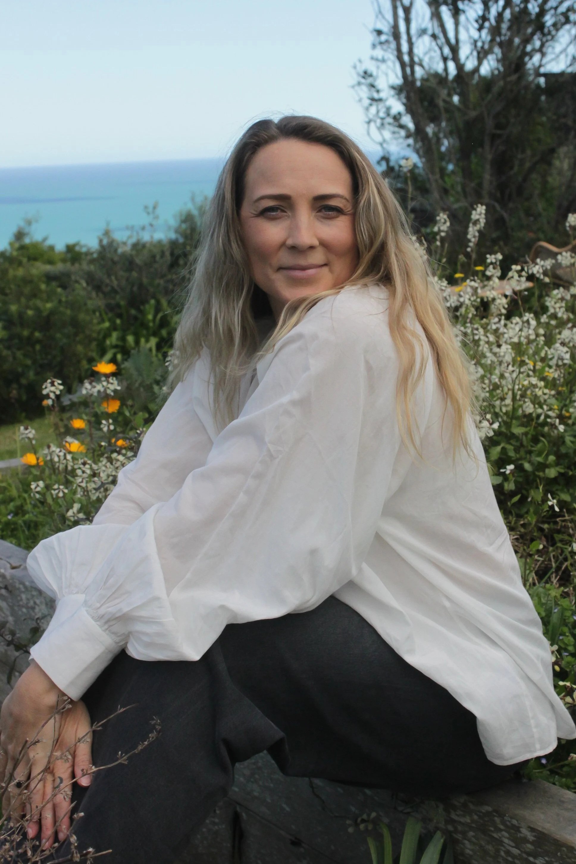A woman with long wavy blonde hair sitting outdoors on a rock, smiling, with a background of trees, wildflowers, and a body of water in the distance.