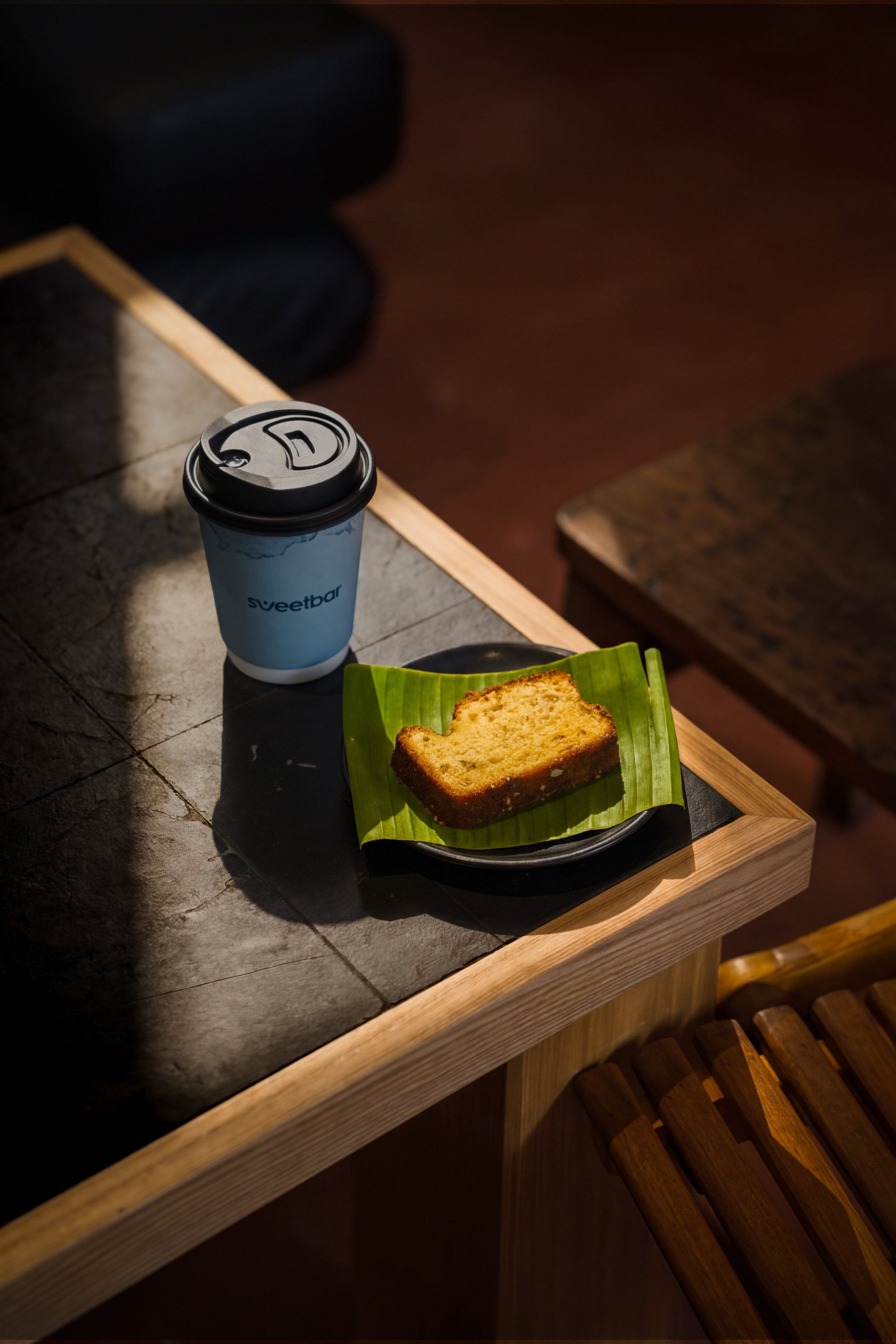 A disposable coffee cup with a black lid and a blue logo that says 'sweetbar' is on a dark table. Next to it is a green paper tray with a slice of banana bread. The setting appears to be on a wooden table in a cozy, dimly lit environment.