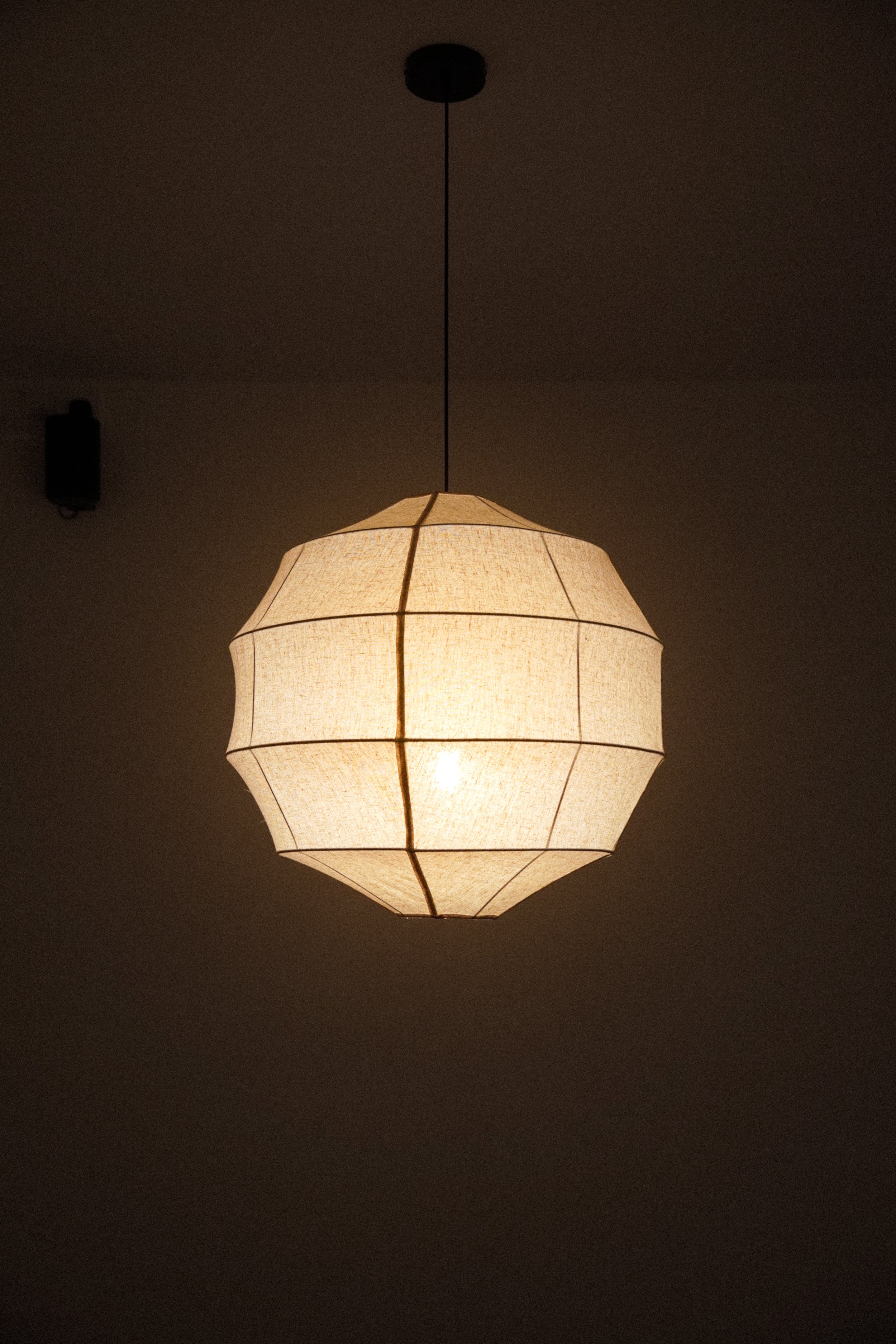 A ceiling lamp with a warm light hangs from a black cord on a plain ceiling, illuminating a dark room.