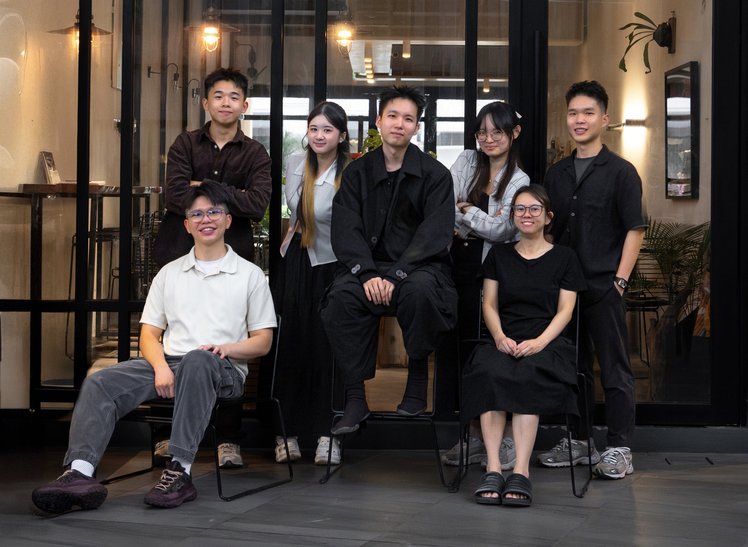 Group of seven young adults posing inside a modern cafe or restaurant with glass wall partitions, warm lighting, and plants.