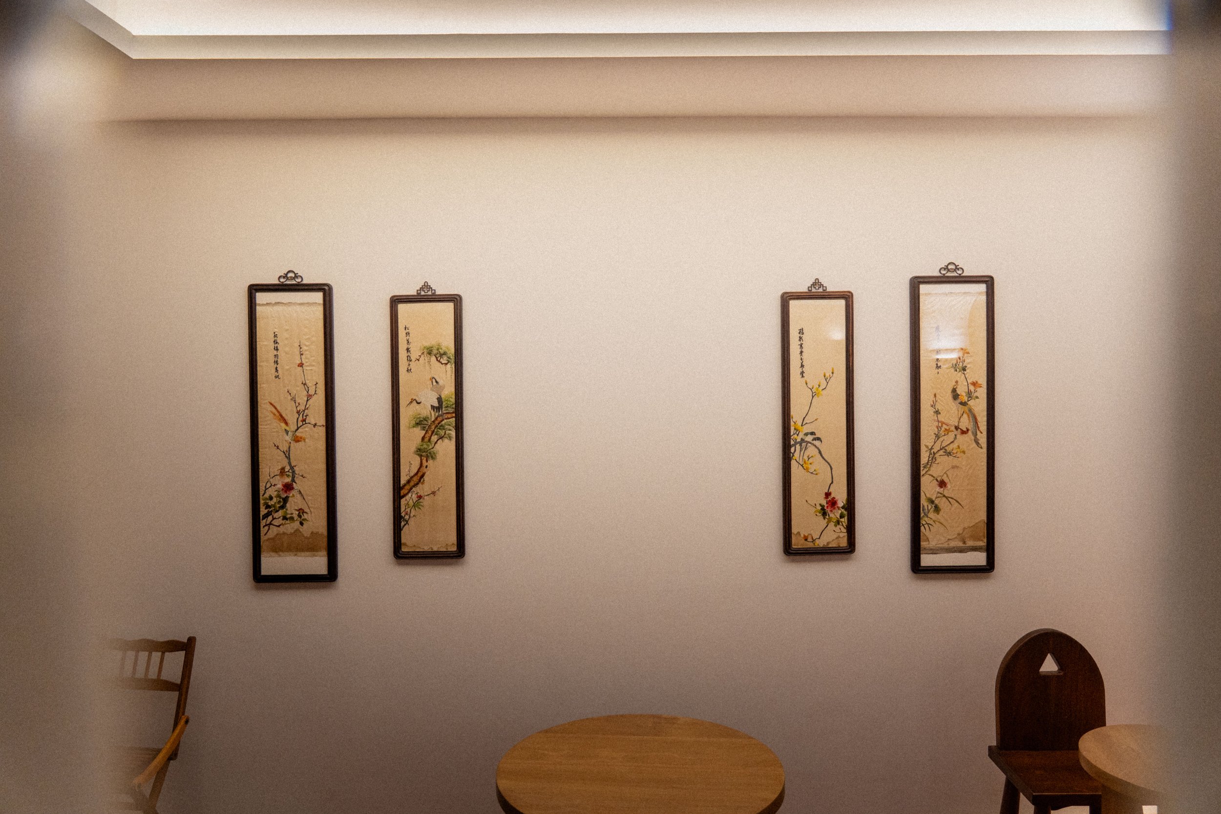 A plain white wall with four framed Asian-style scroll paintings, each featuring traditional Japanese or Chinese artwork and calligraphy, hanging vertically in a row. Part of a wooden chair and a table are visible in the foreground.