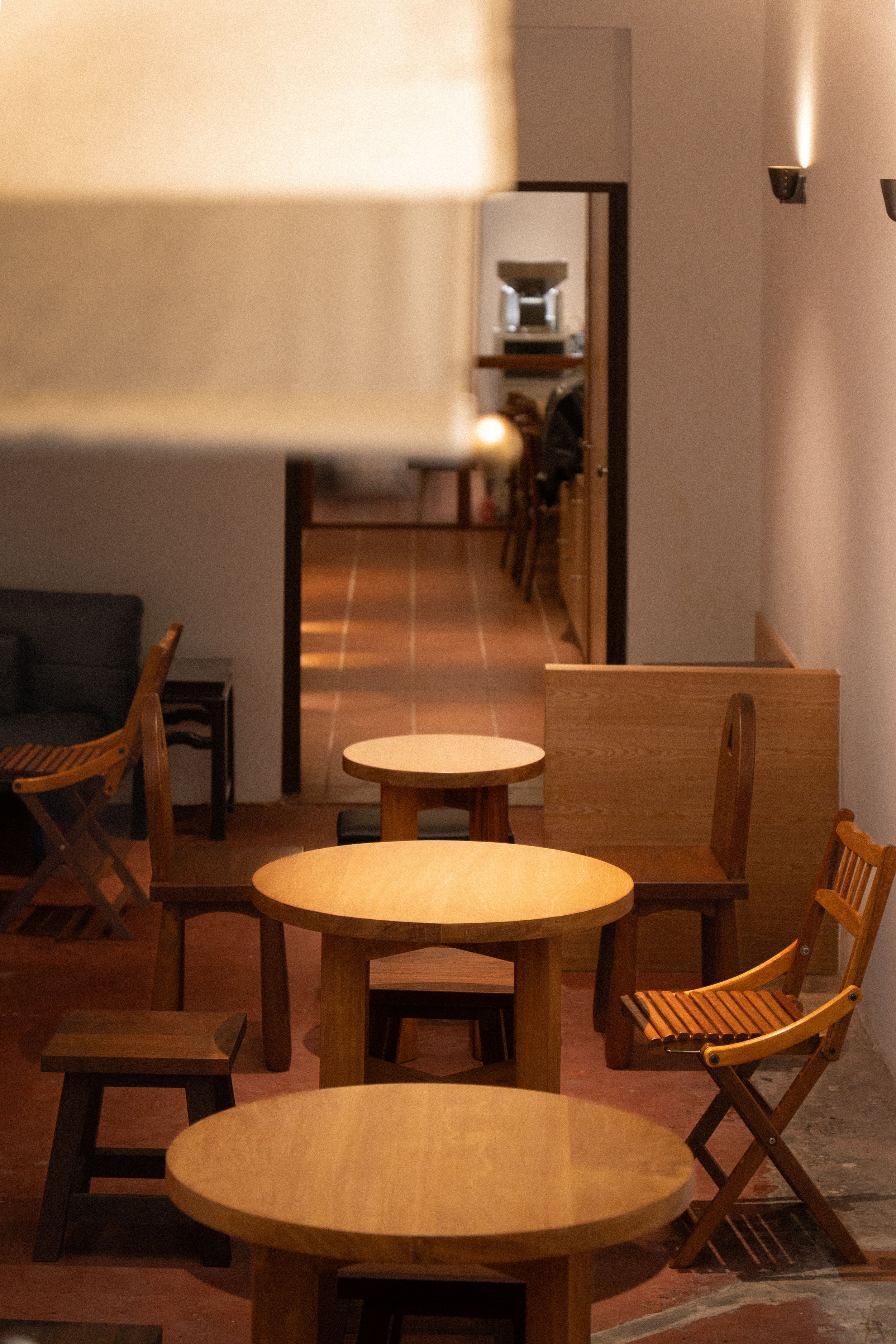 Dining area with multiple small round wooden tables and wooden chairs, a partial view of a black couch, and a hallway with terracotta tiled floor leading to a room with a mirror and a shelf in the background.