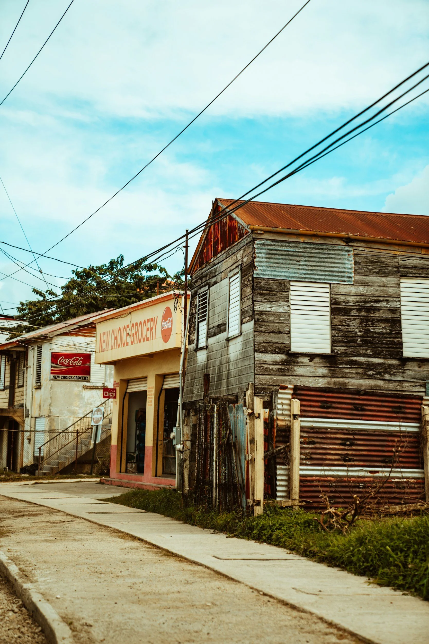 An old weathered building with a rusty metal roof and wood siding, next to a newer convenience store with signs displaying 'New Choice Grocery' and Coca-Cola branding. The street in front is a concrete sidewalk with grass on the side, under a partly cloudy sky.
