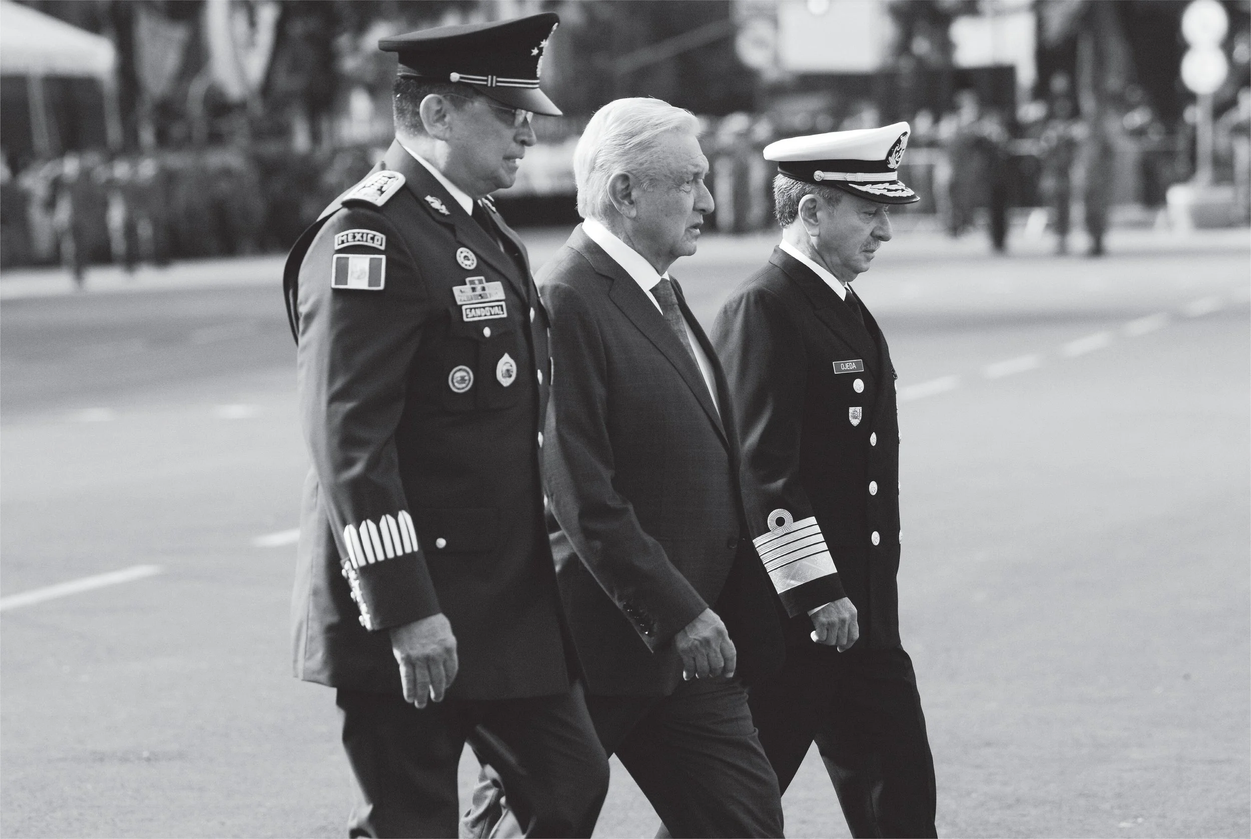 Three men dressed in formal uniforms and a suit are walking outdoors in a line, with solemn expressions. One man is wearing a military uniform with various patches, another is in a suit, and the third is wearing a naval uniform with a white cap.