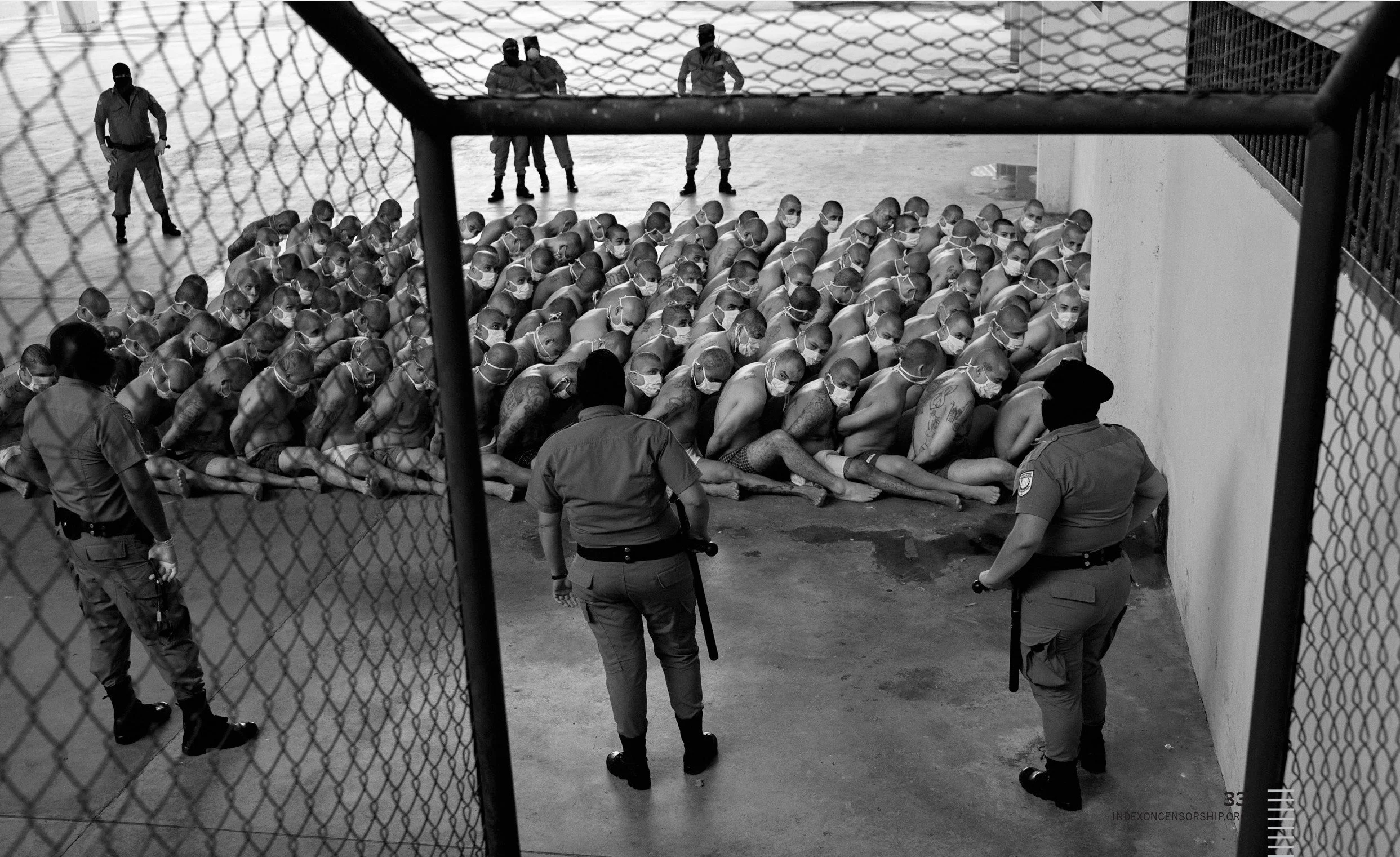 A group of prisoners, all shirtless and wearing face masks, kneeling outside in a line, with six guards standing around them. The scene appears to be at a detention facility, viewed through a chain-link fence.