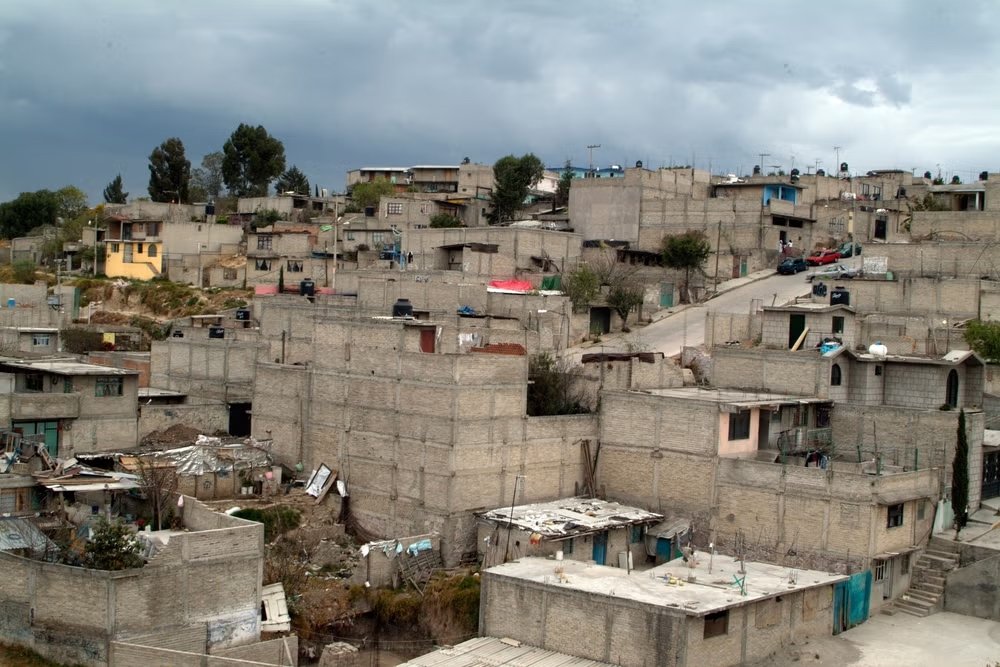 A hillside neighborhood with numerous unfinished concrete block houses, some with small trees and cars visible, under a cloudy sky.