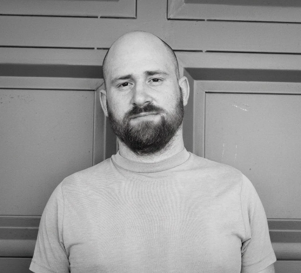 Black and white photo of a man with a beard and mustache, wearing a light-colored T-shirt, standing against a paneled wall background.