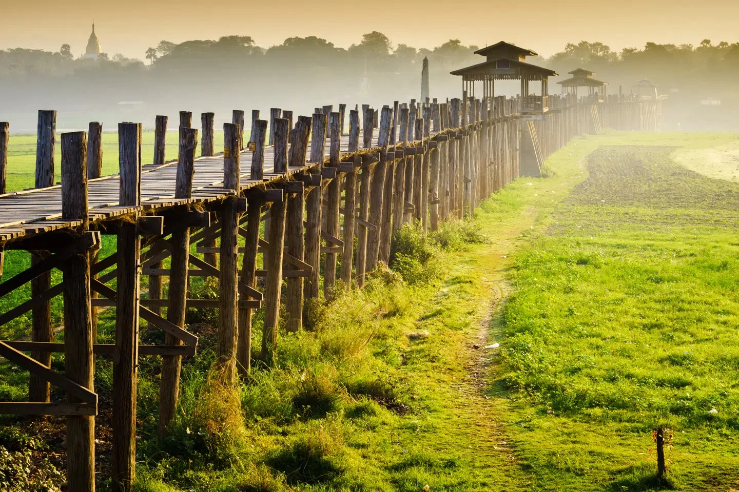 A wooden bridge with small pavilions stretches over a green field during sunrise or sunset.