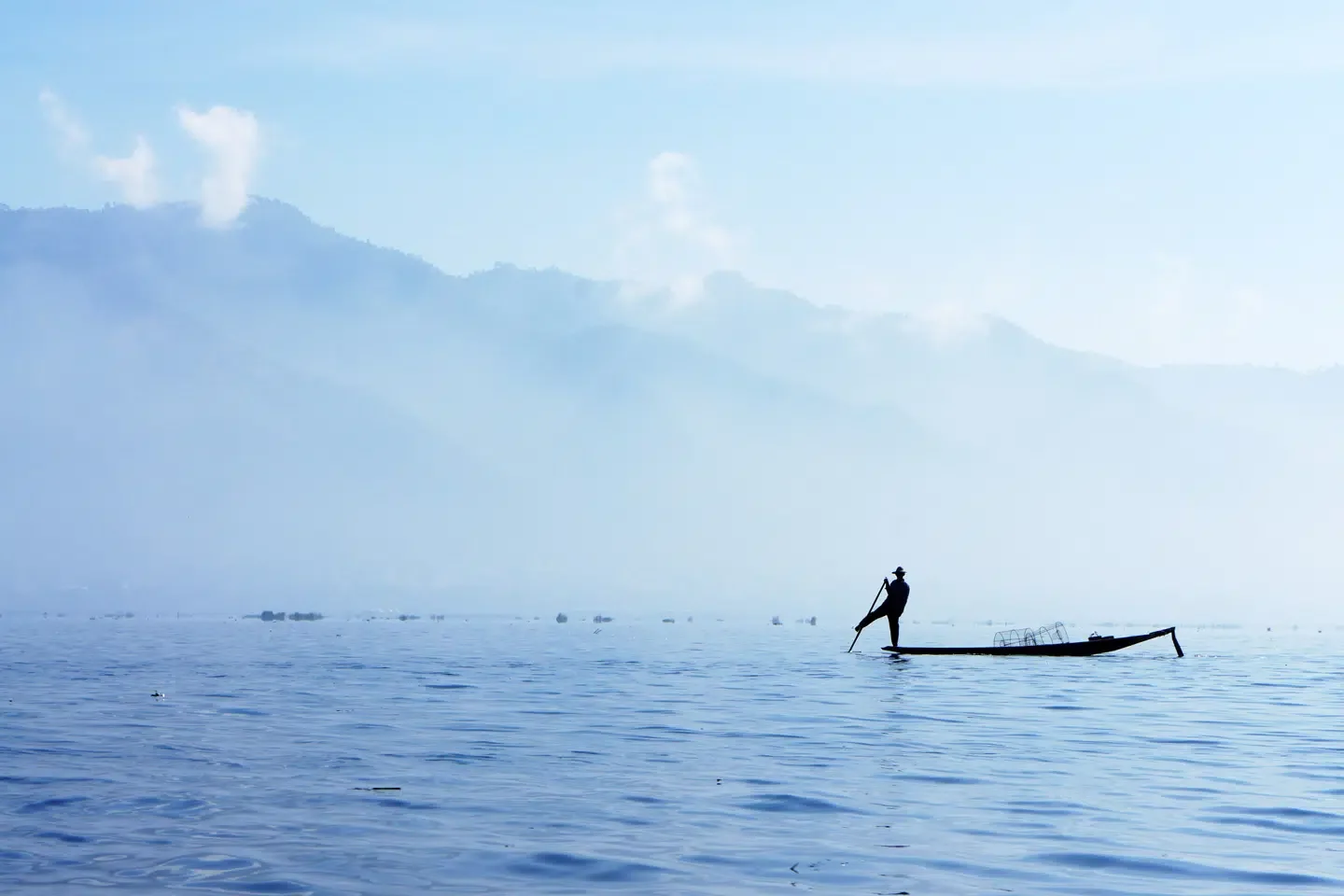 Silhouette of a person standing on a small boat or raft on a calm body of water with mountains and sky in the background.