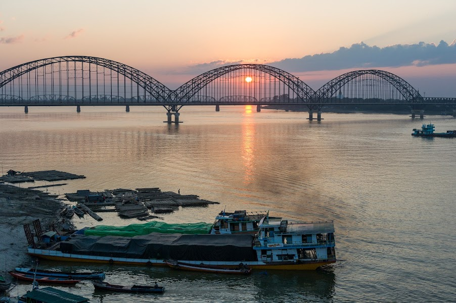 Sunset over a large river with a bridge in the background and boats docked along the shore in the foreground.