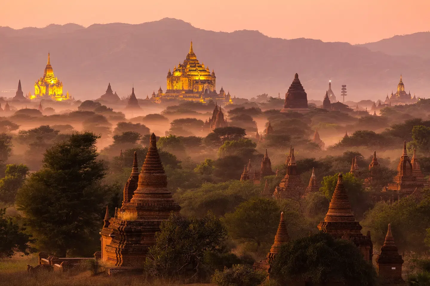 Several ancient temples and pagodas in a misty landscape during sunrise or sunset, with mountain ranges in the background.