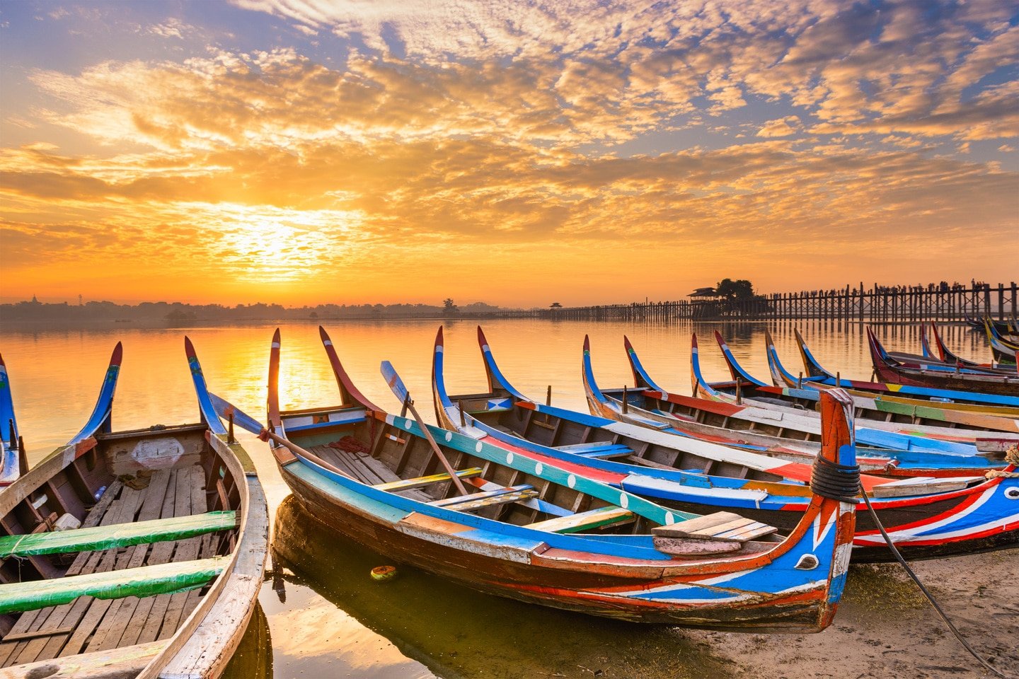 Colorful traditional wooden boats docked along a riverbank during a vibrant sunset with a bridge and trees in the background.