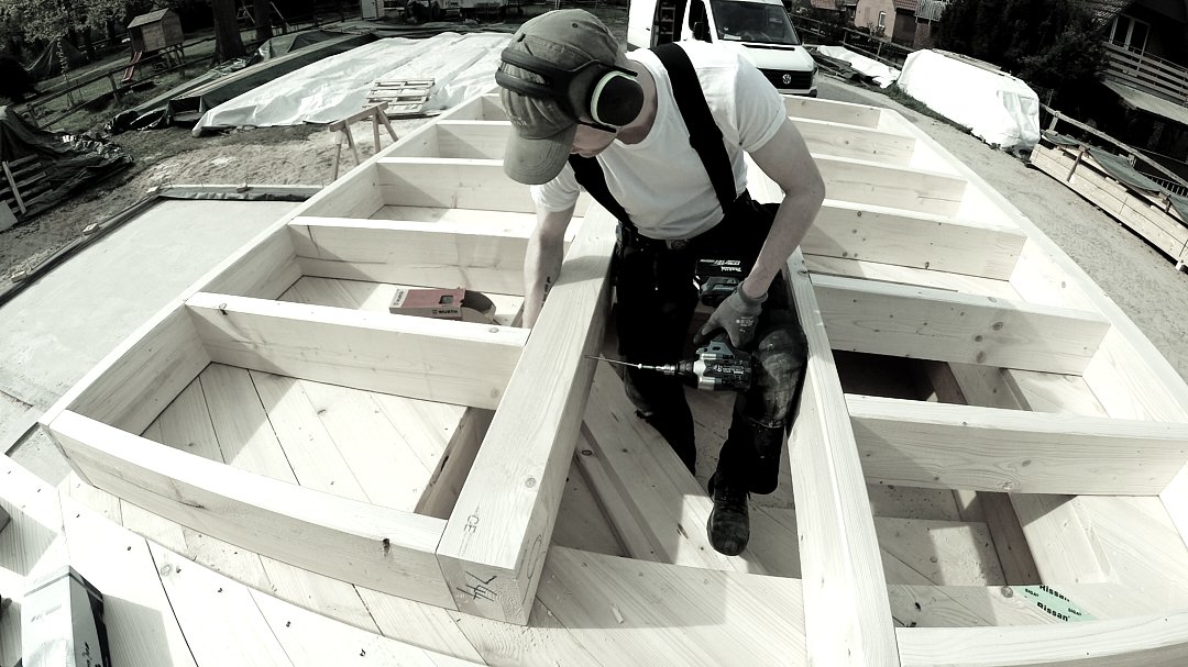 Ein Handwerker arbeitet auf einem Baugerüst und befestigt Holzbalken mit einem Bohrschrauber.