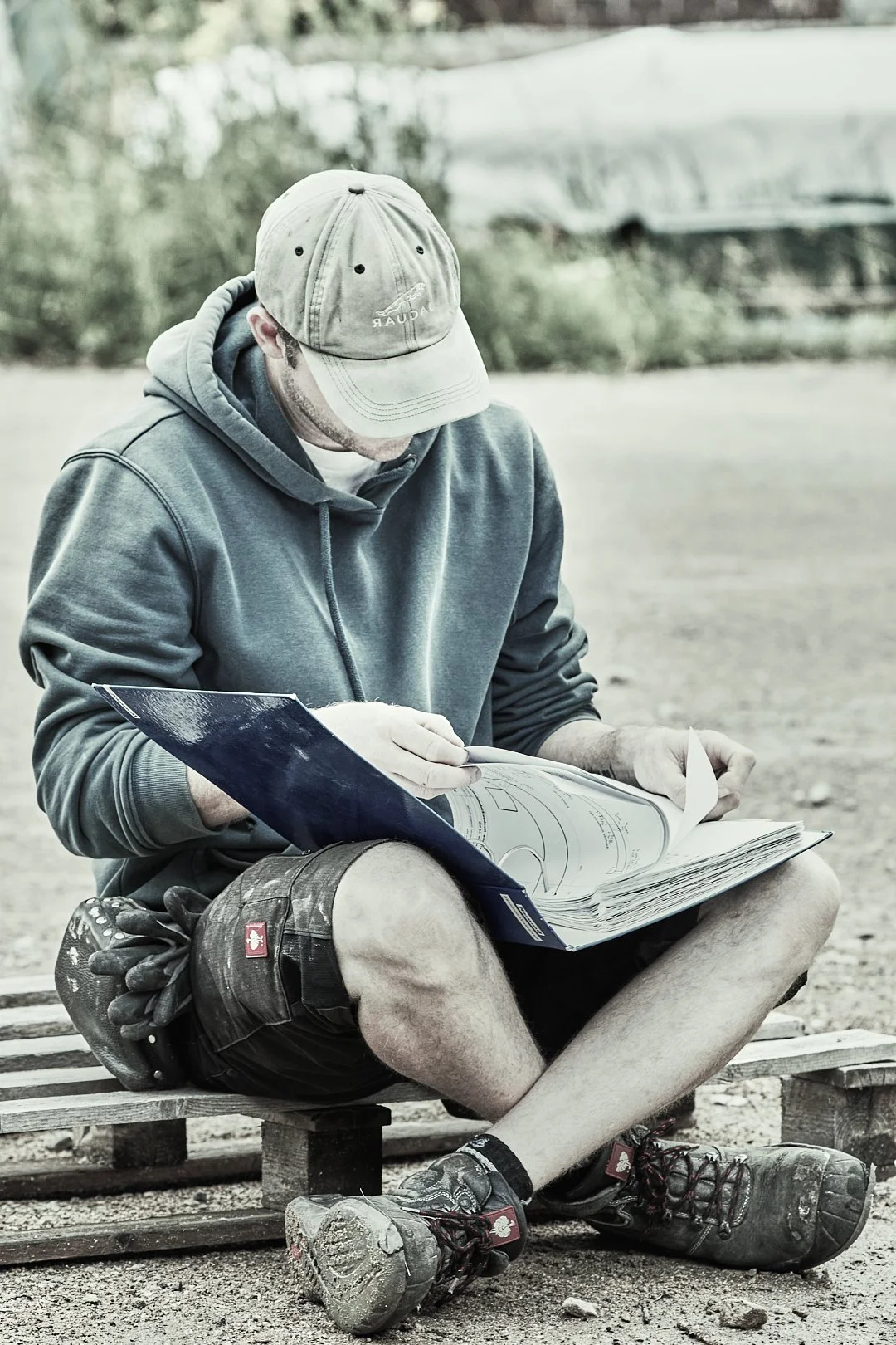 Ein Mann sitzt auf einem Holzbänkchen im Freien, trägt ein graues Hoodie, eine beige Mütze und erkundet einen großen Plan oder eine Zeichnung.