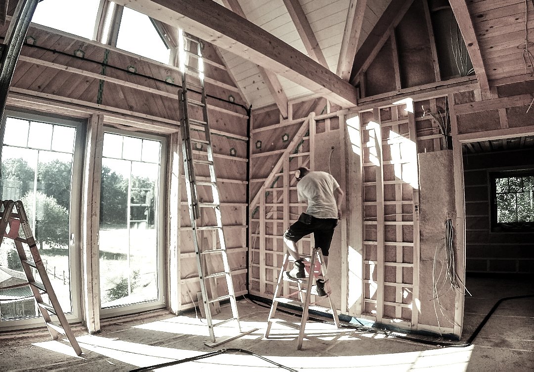 Mann bei der Arbeit im Rohbau eines Hauses, während er auf einer Leiter steht, mit Holzrahmen und -wänden; große Fenster lassen Tageslicht in den Raum.