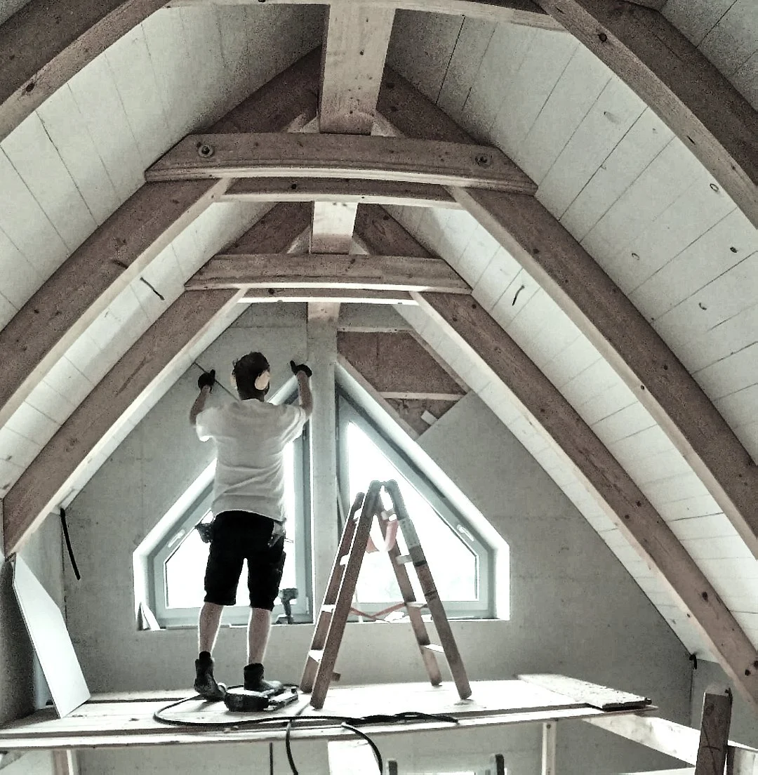 Mensch arbeitet in einem Dachgeschoss beim Renovieren, steht auf einer Leiter, bei einem Fenster, mit Holzstützen und Baumaterialien im Raum.