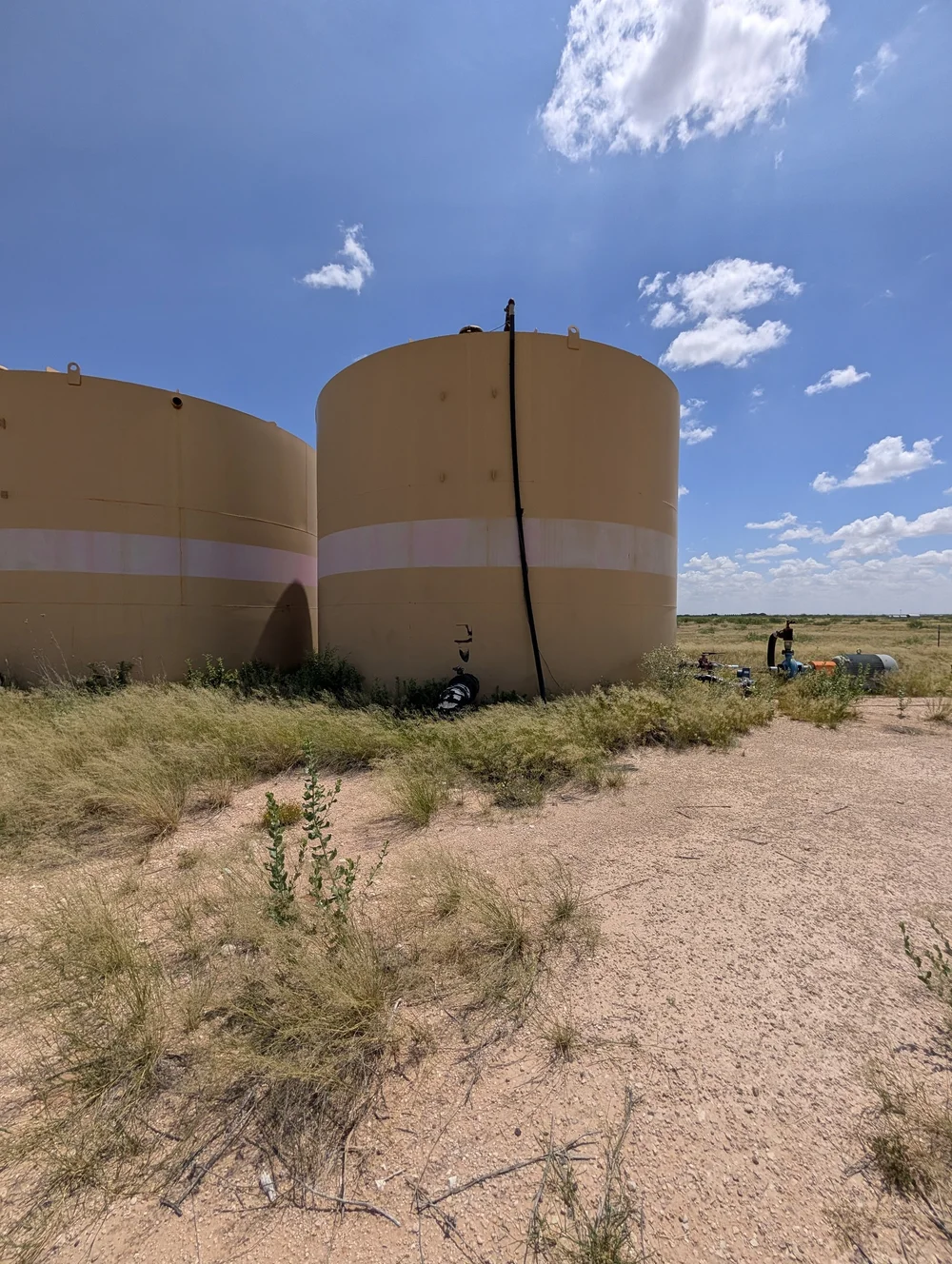 Two storage tanks with grass and shrubs growing against the base and chime, and pipework at ground level