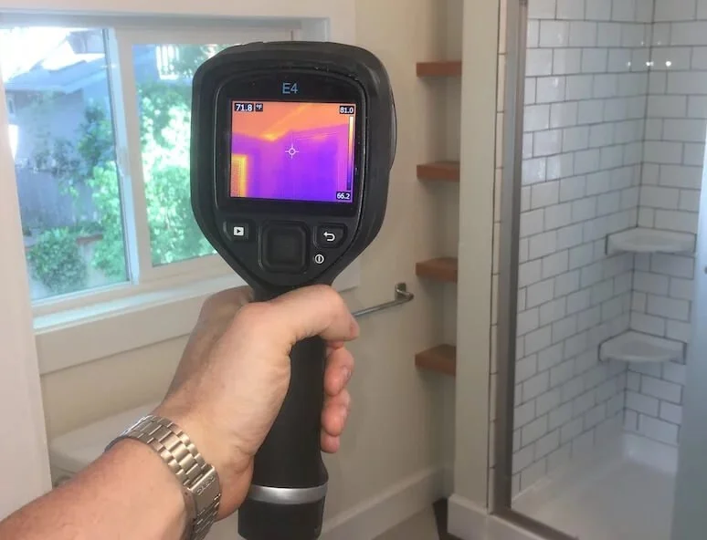 Person holding a thermal imaging camera in a bathroom with a shower and window.