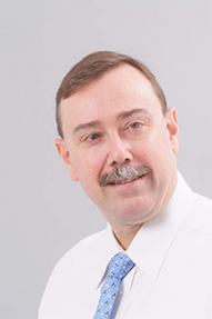 Close-up of a middle-aged man with brown hair, mustache, wearing a white shirt and blue polka dot tie, smiling against a light gray background.