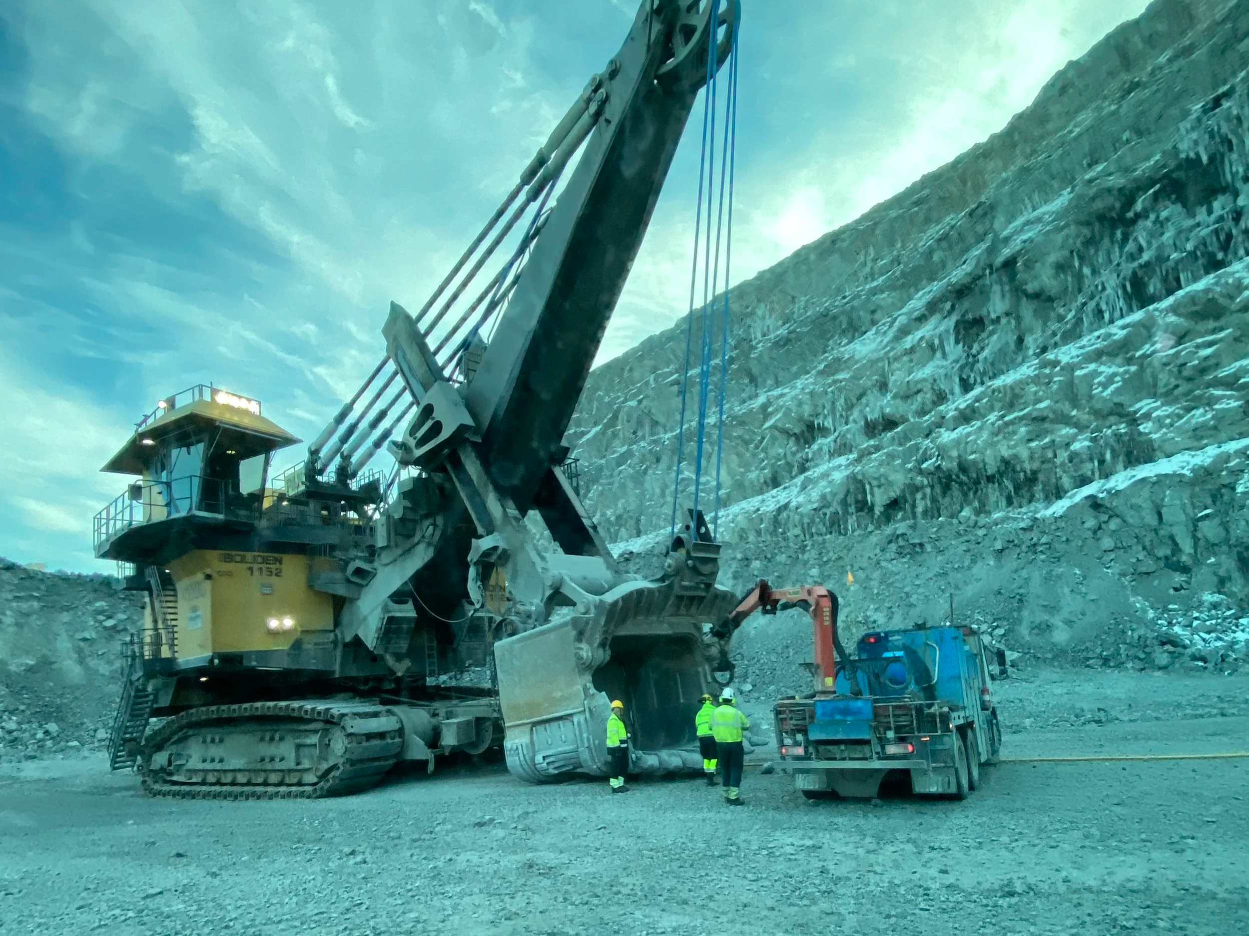 A large yellow construction excavator working in a rocky, icy environment with workers in safety gear nearby.