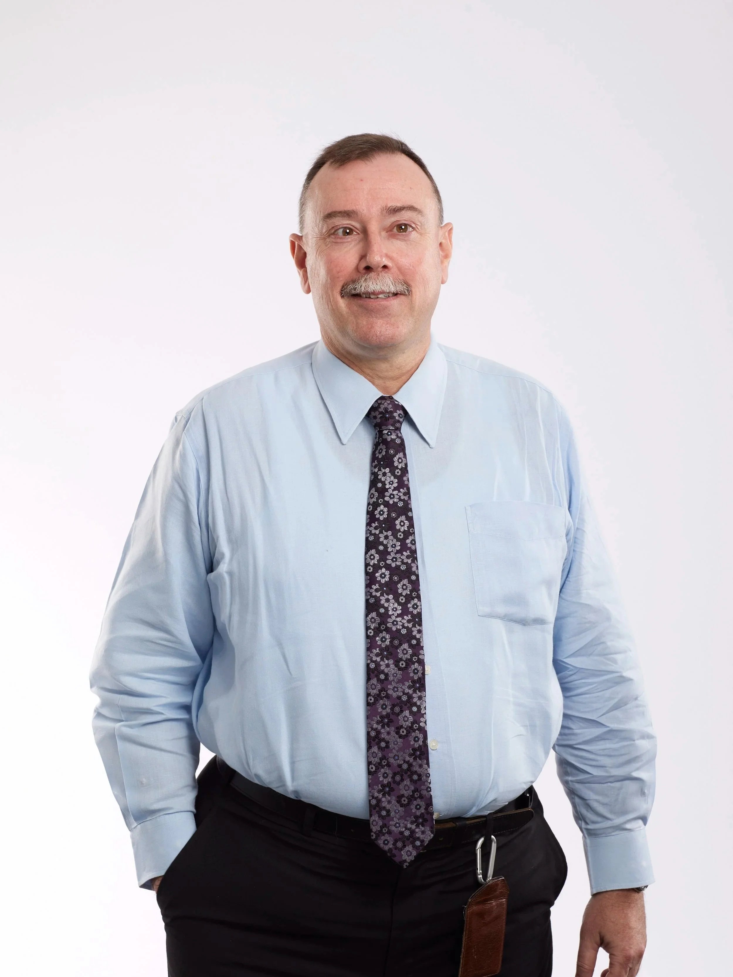 A middle-aged man with short brown hair, a mustache, and light skin, wearing a light blue dress shirt, a dark floral tie, and black pants, standing against a plain white background.