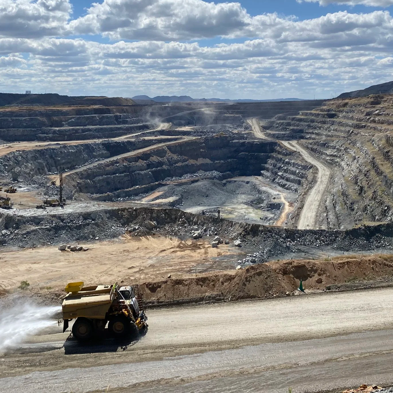 Open-pit mining operation with a large dump truck driving on a dusty road in the foreground, and terraced rocky slopes with dust clouds and machinery in the background under a partly cloudy sky.