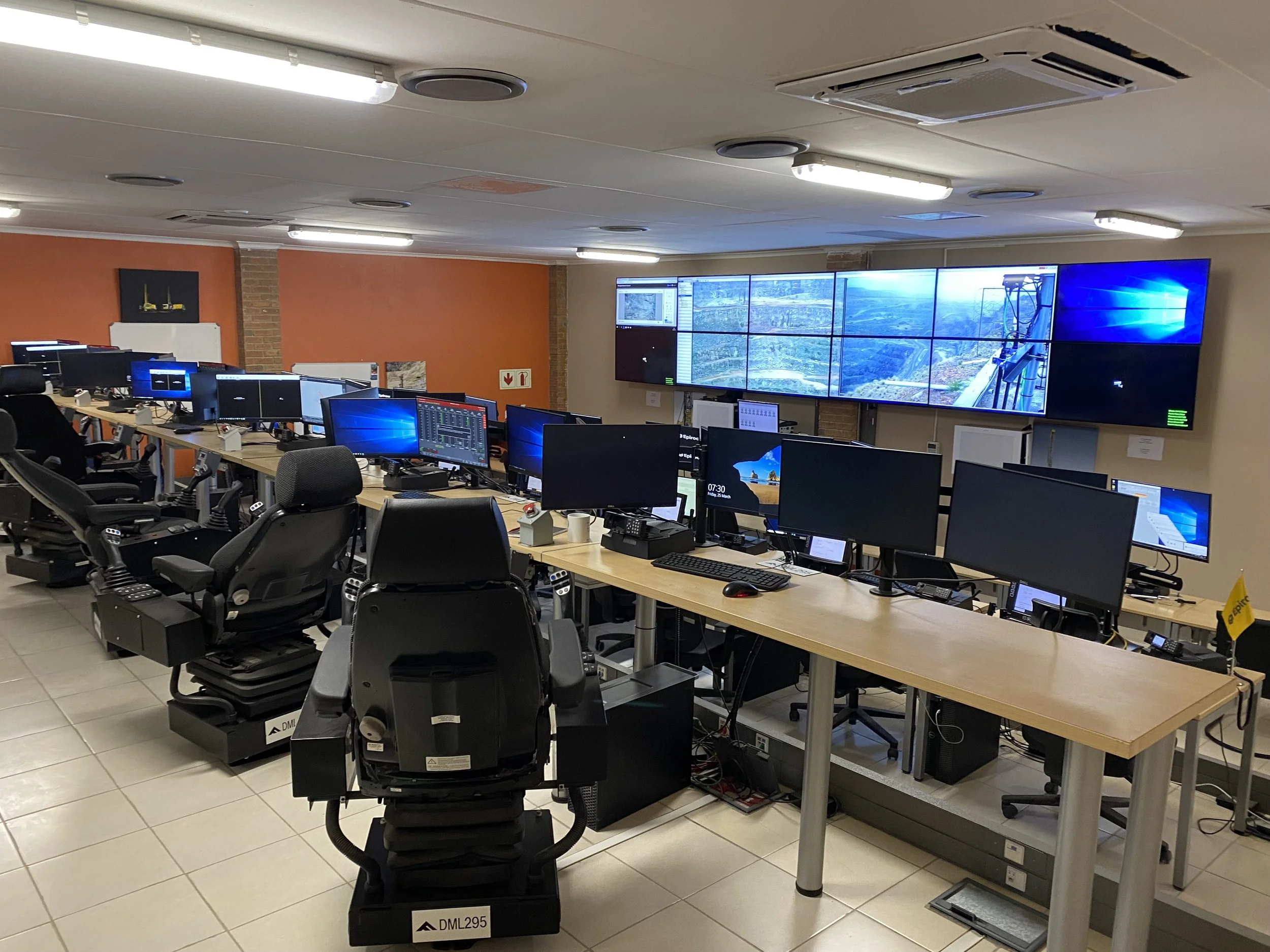 An office control room with multiple computer monitors and large video screens on the wall displaying outdoor and technical views. Rows of control desks and chairs are visible.