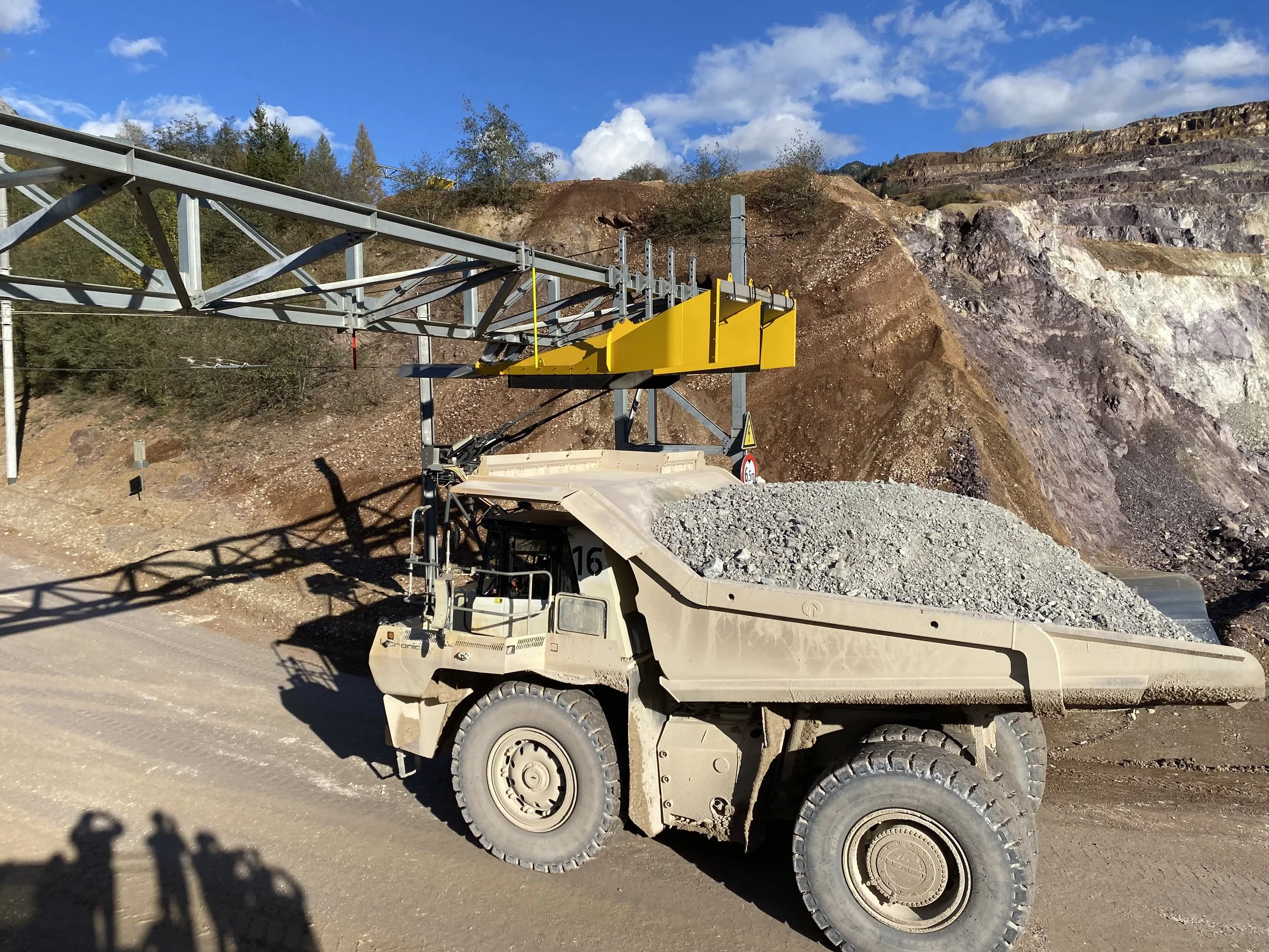 A construction vehicle with a crane arm and a yellow attachment working on a rocky hillside in a mountainous area under a partly cloudy sky.