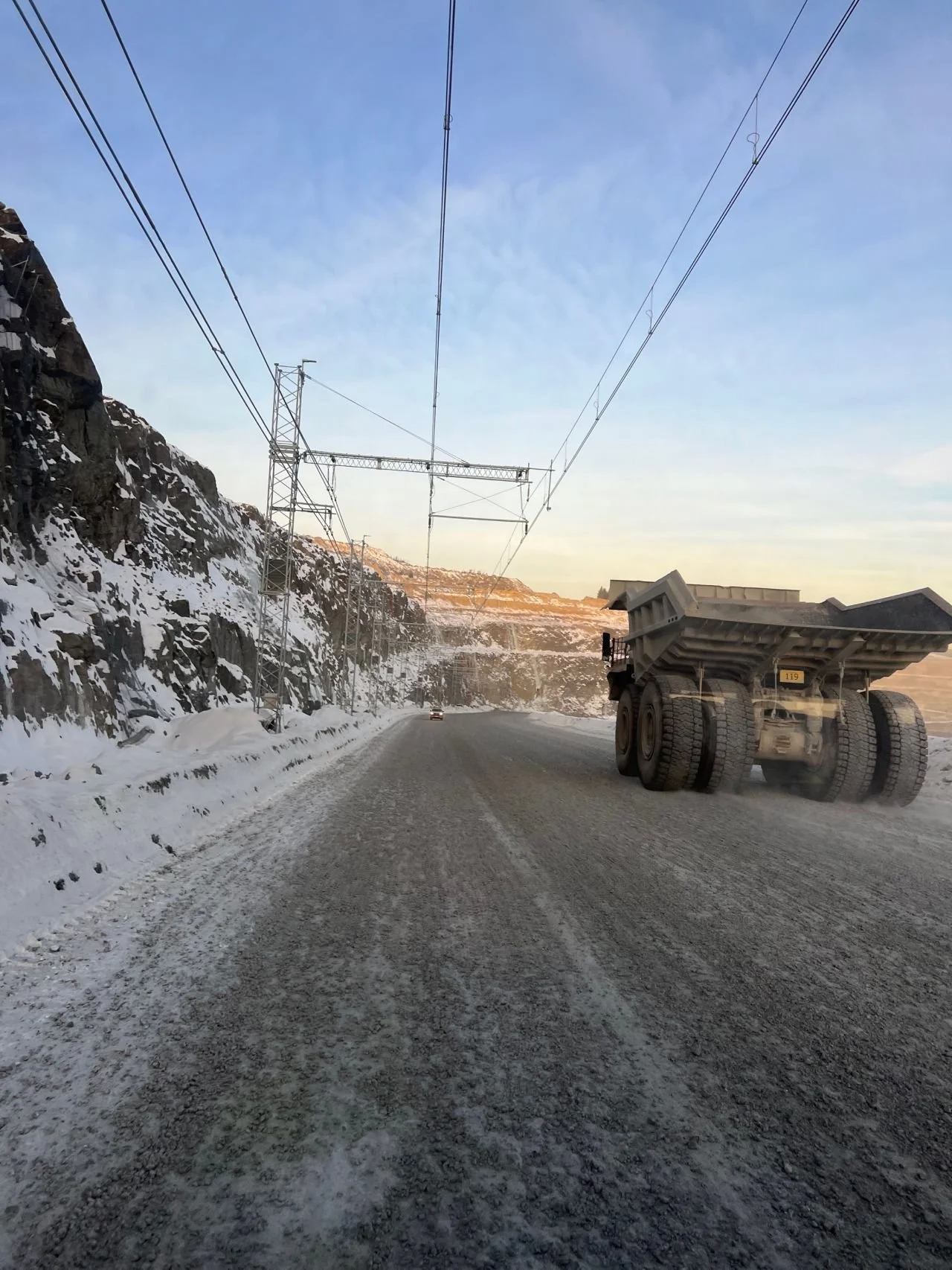 A snow-covered mountain road with a large dump truck and a vehicle in the distance. Power lines and towers run alongside the road, with snow and ice on the ground and mountains in the background.