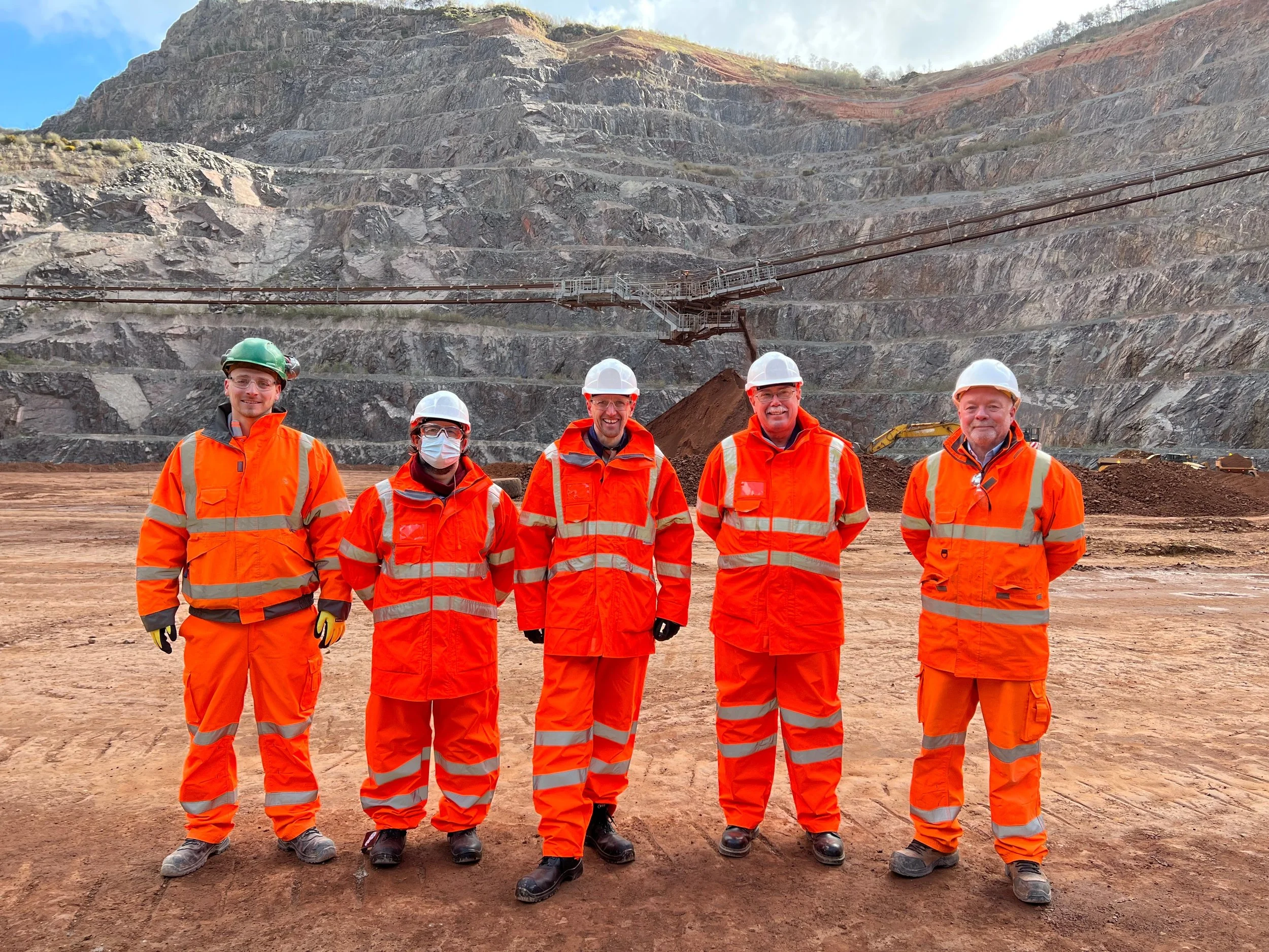 Six workers in orange safety uniforms and white helmets standing in front of a hilly, rocky landscape with an industrial mining structure in the background.