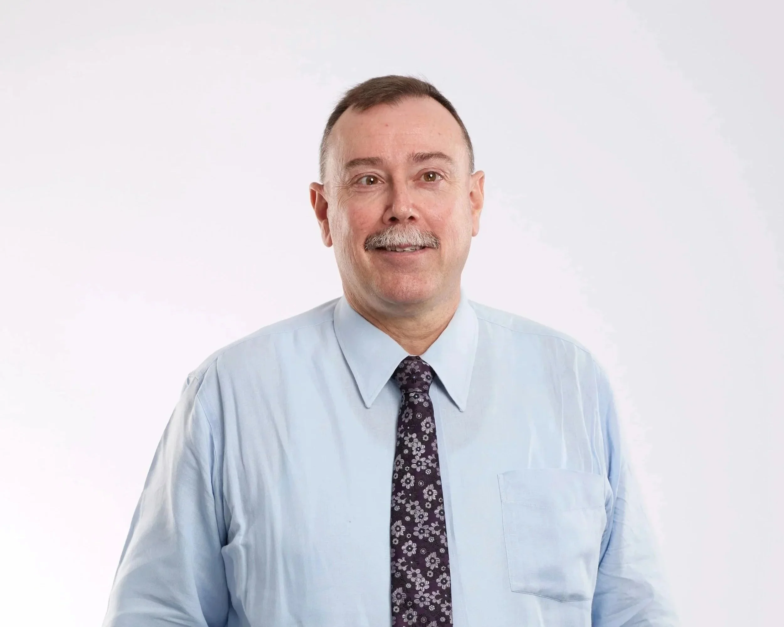 A middle-aged man with short brown hair, a mustache, wearing a light blue dress shirt and a dark floral tie, standing against a plain white background.